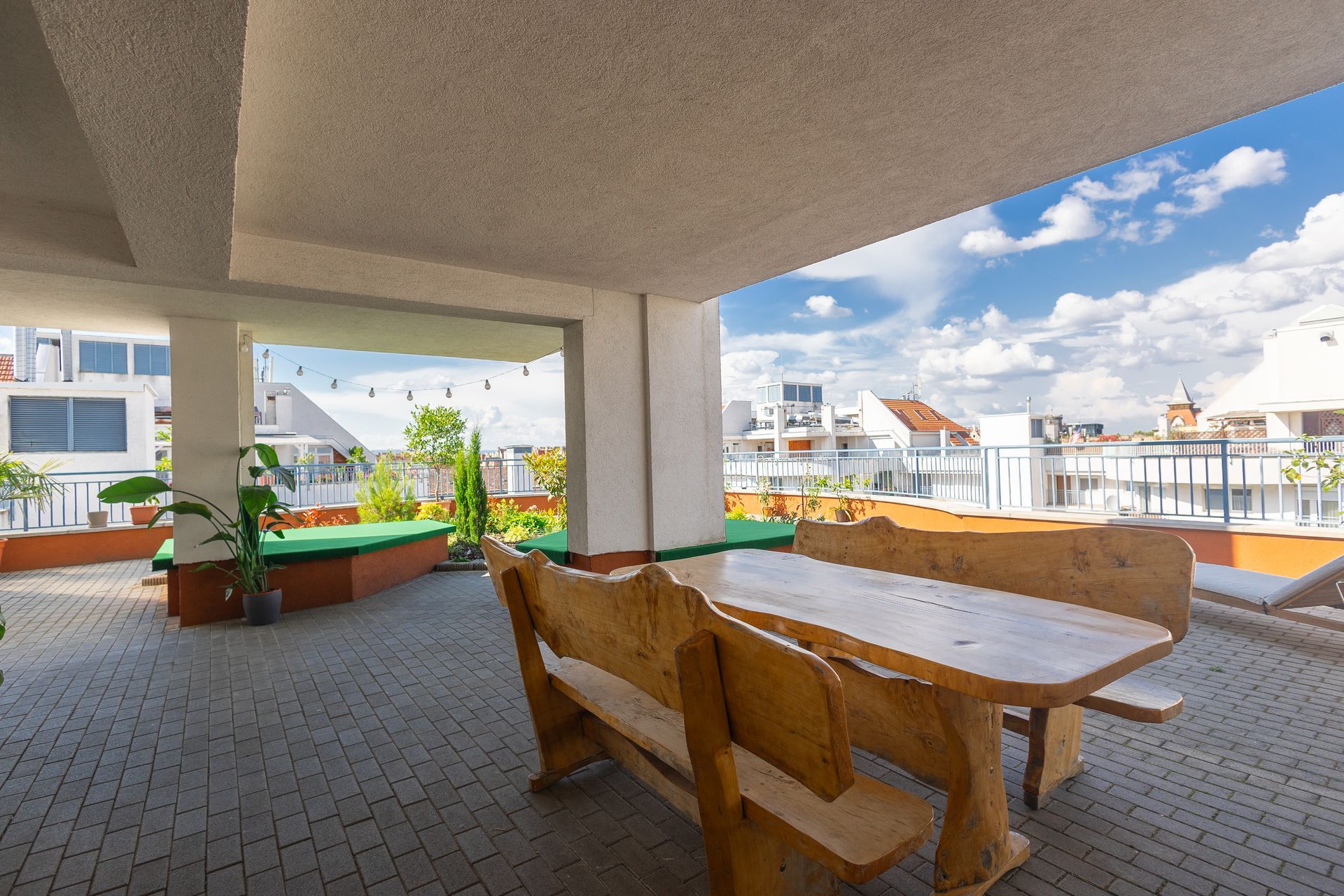 Covered outdoor patio with wooden table and benches, overlooking a city on a sunny day.