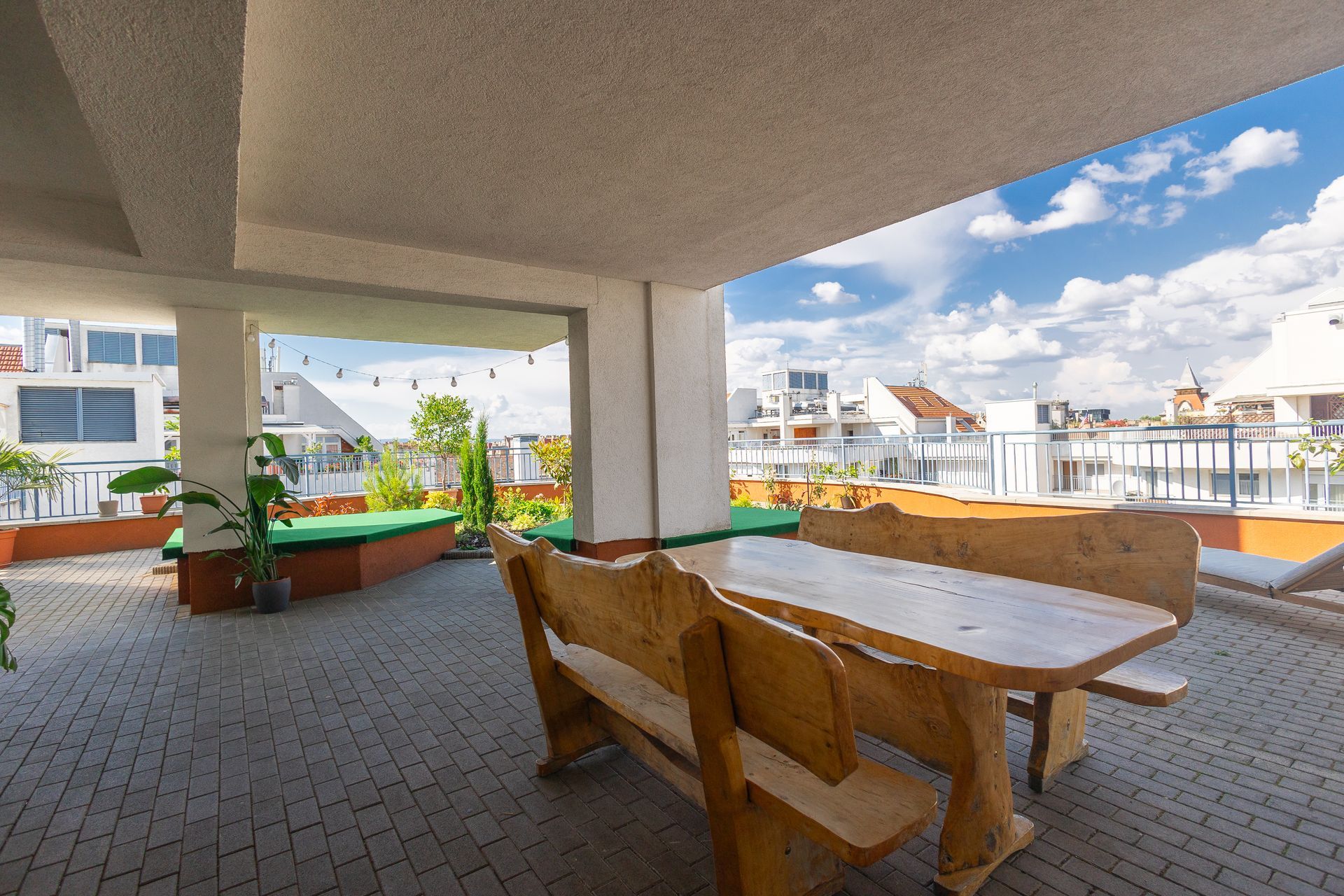 Outdoor patio with a wooden table and benches, city view, blue sky with clouds.