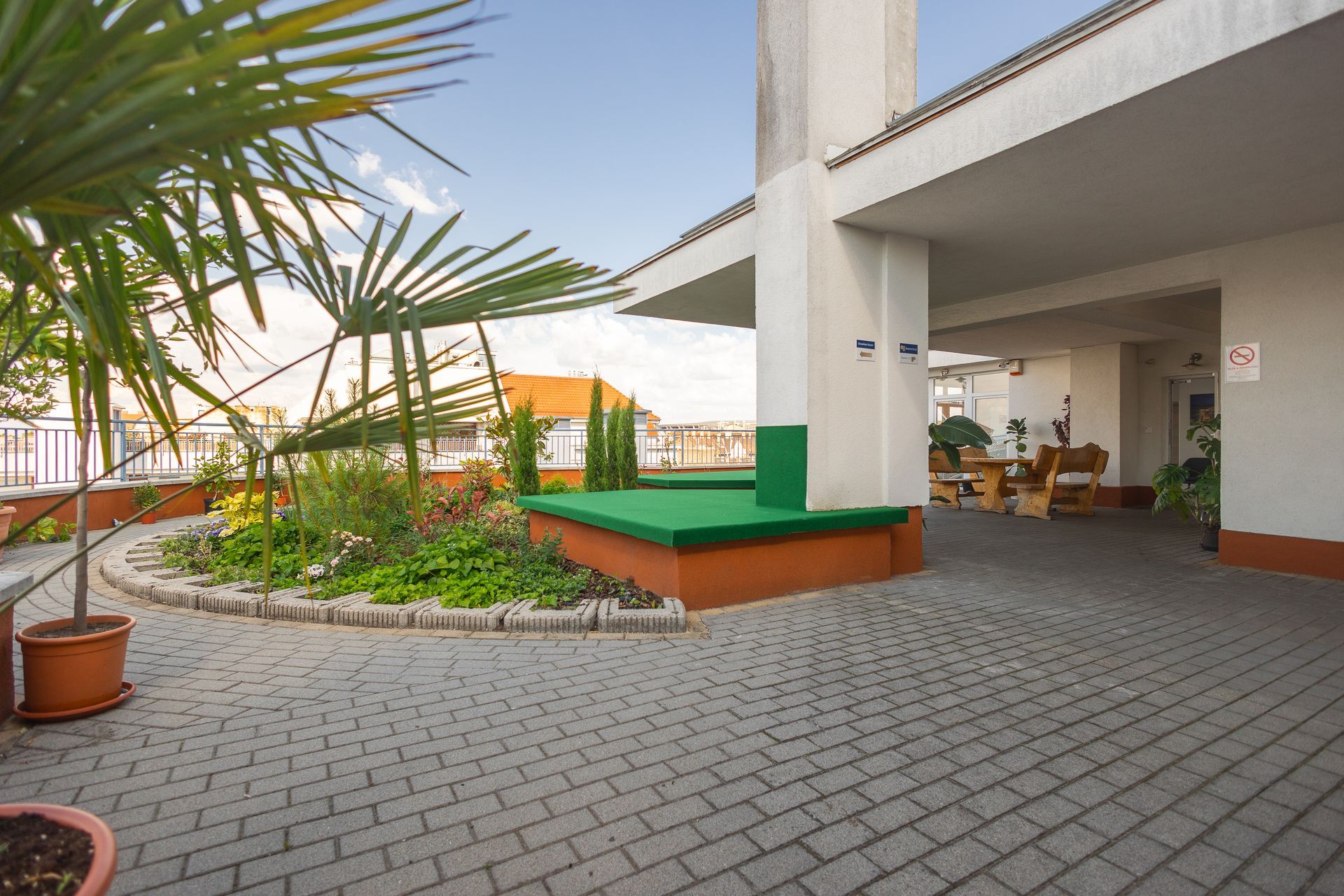 Terrace with brick pavers, flowerbeds, and seating under a covered area, blue sky.