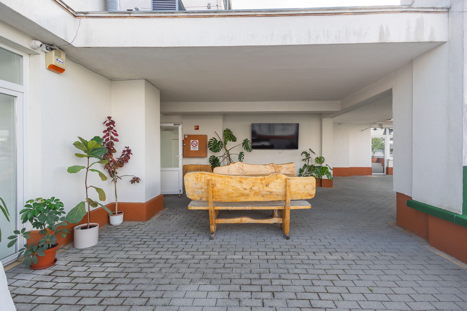 Covered outdoor seating area with a wooden bench, plants, and a mounted TV on a white wall.