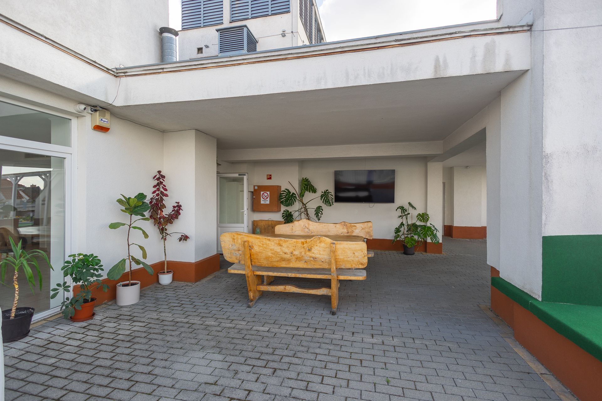 Covered patio with wooden benches, plants, and a wall-mounted TV. Paved flooring, white walls with red and green accents.