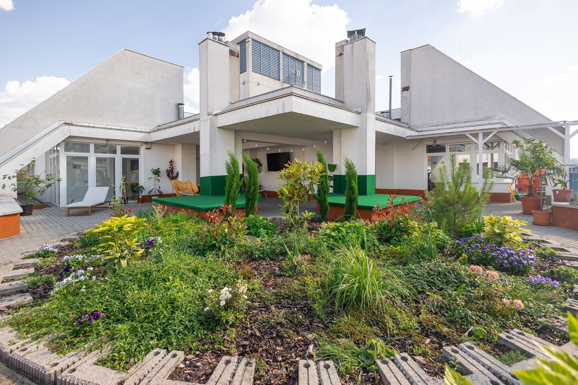 A white building with a garden. A porch, and rooftop with chimneys, plants, and pots.