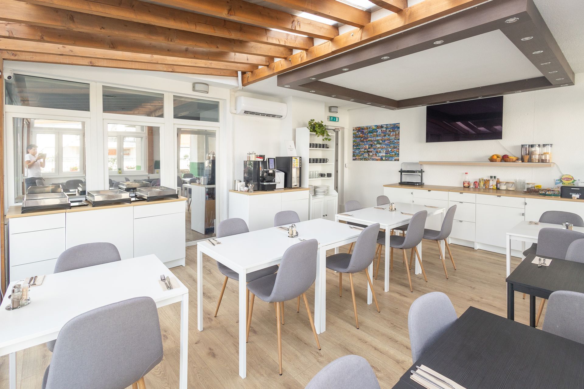 Dining room with white tables, gray chairs, and coffee station. Wooden ceiling and light wood floors.