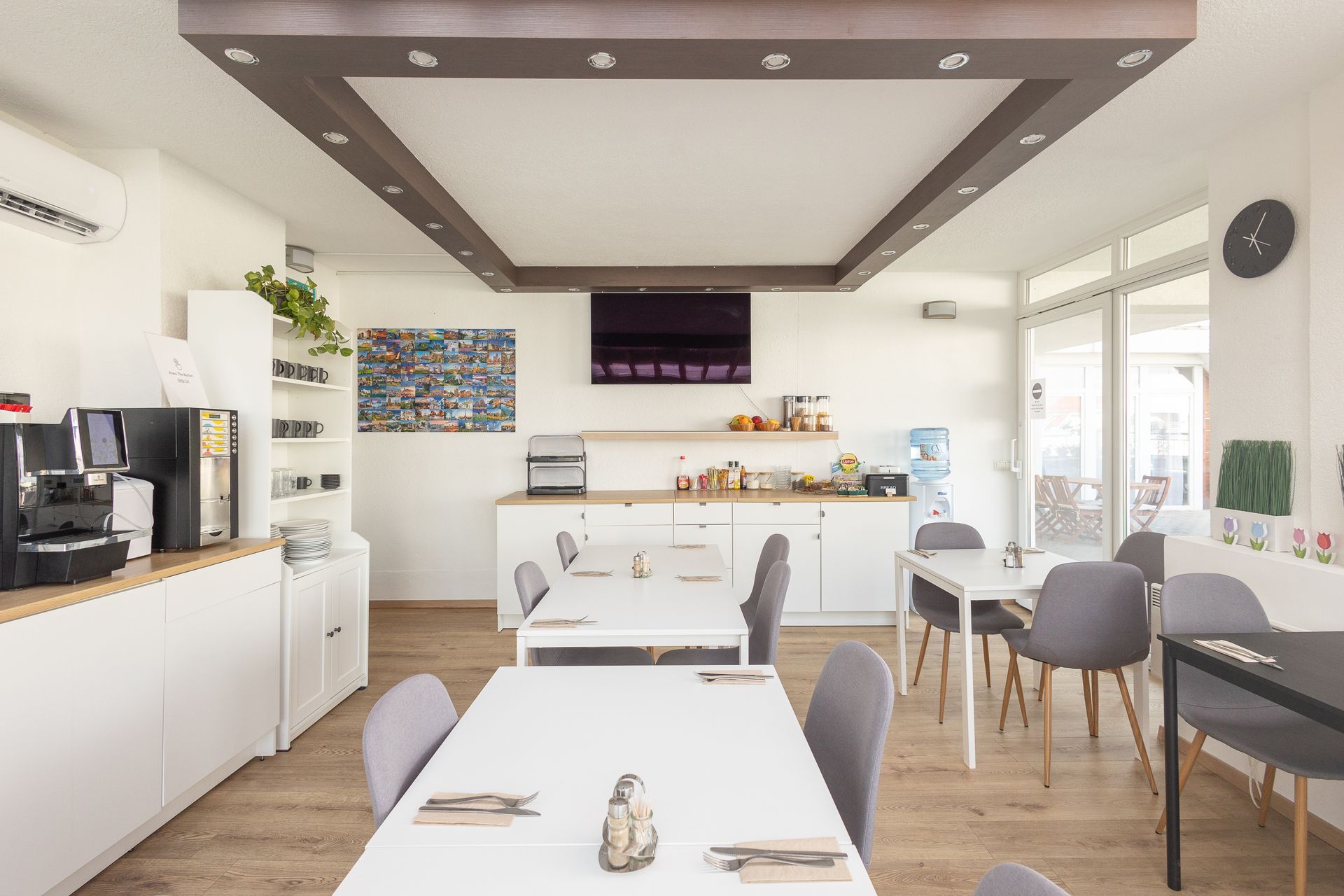 Bright dining area with white cabinets, tables, and ceiling; wooden floor. Includes a coffee machine, TV, and gray chairs.