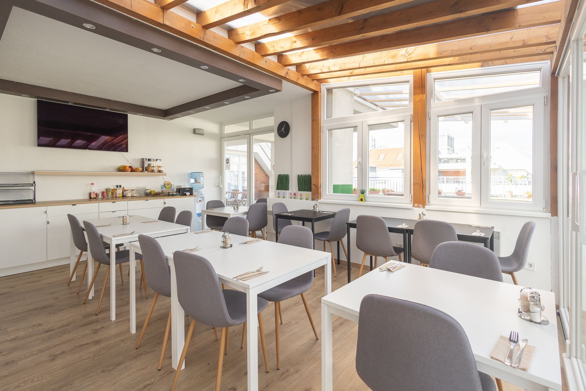 A bright dining area with white tables and grey chairs. Windows and a TV are visible.