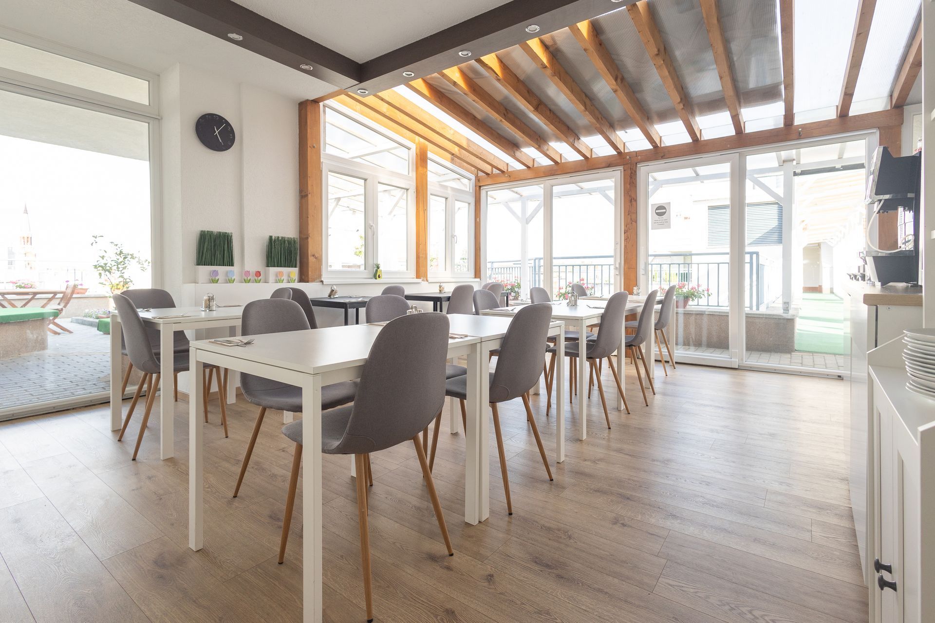 Dining room with white tables, gray chairs, and large windows. Wooden ceiling beams and light wood floors.