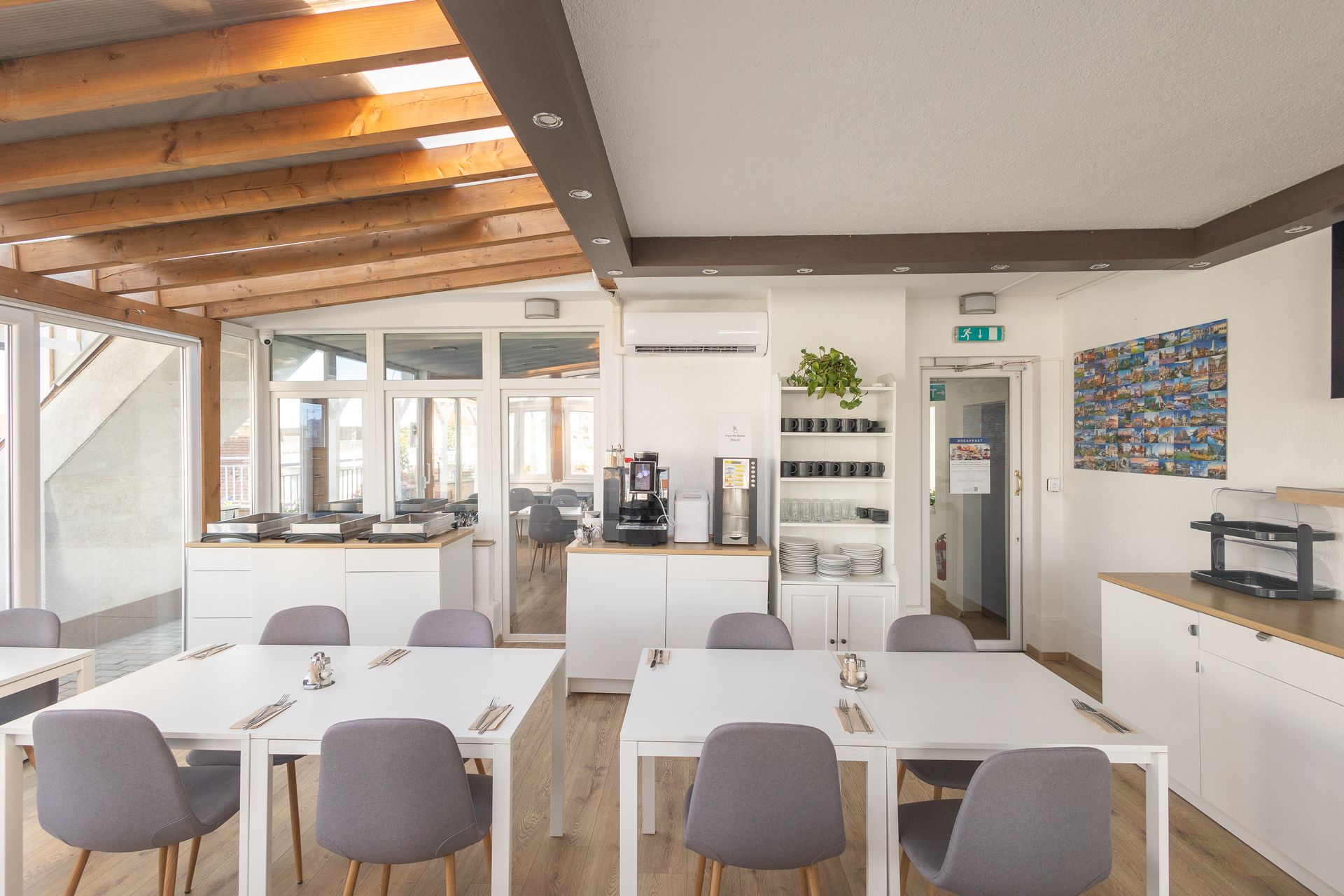 Bright dining area with white tables, gray chairs, coffee station, and a wooden beam ceiling.