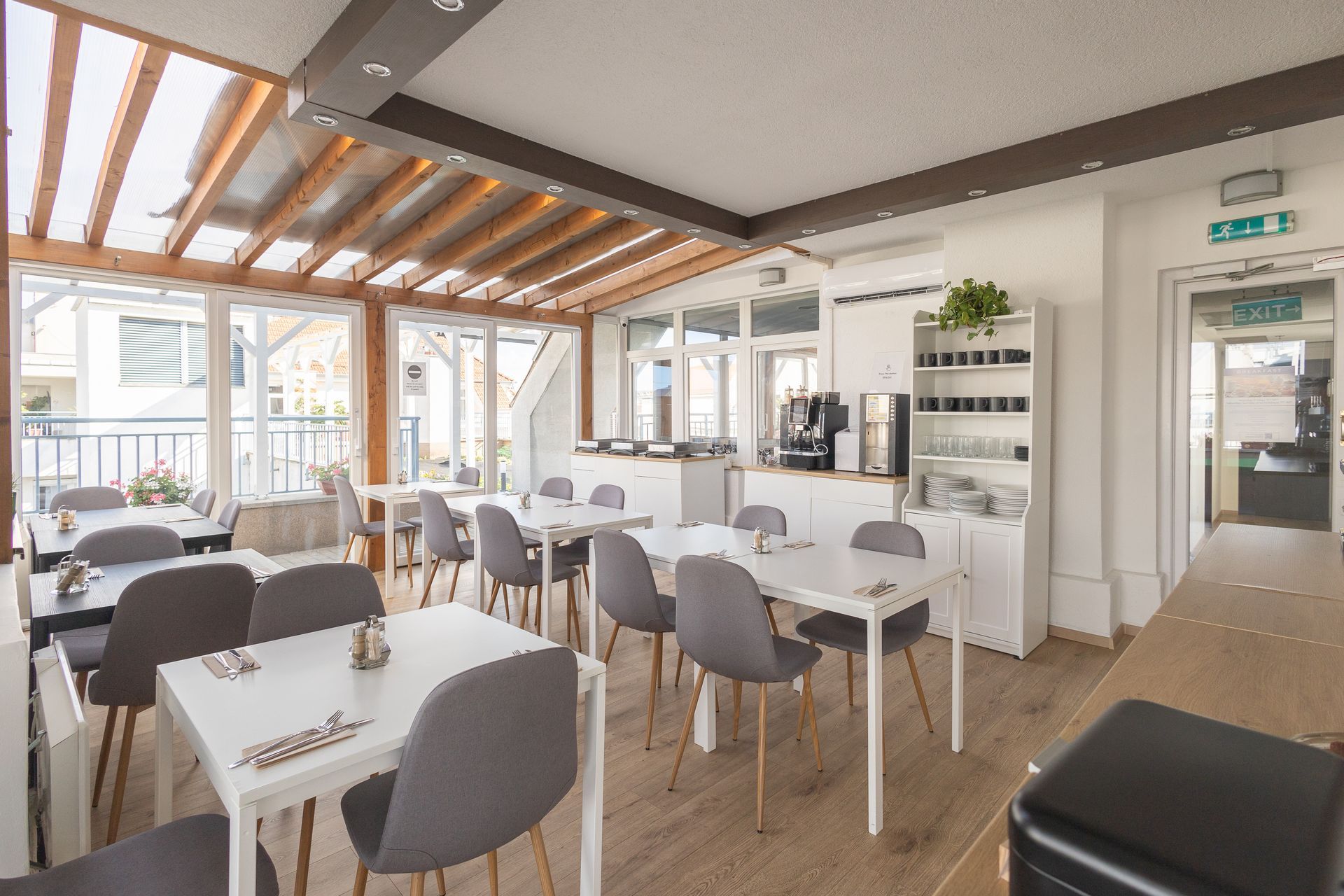 Bright cafe with white tables and grey chairs under a wooden beamed ceiling, natural light coming through a glass paneled wall.