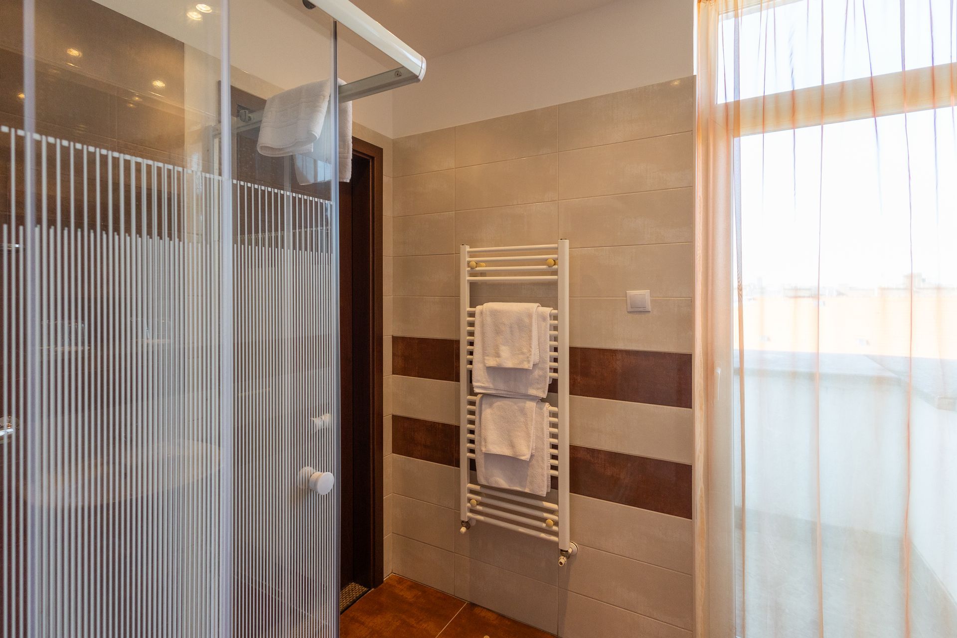 Bathroom with glass shower, towel warmer, beige tile, and a window with sheer curtains.