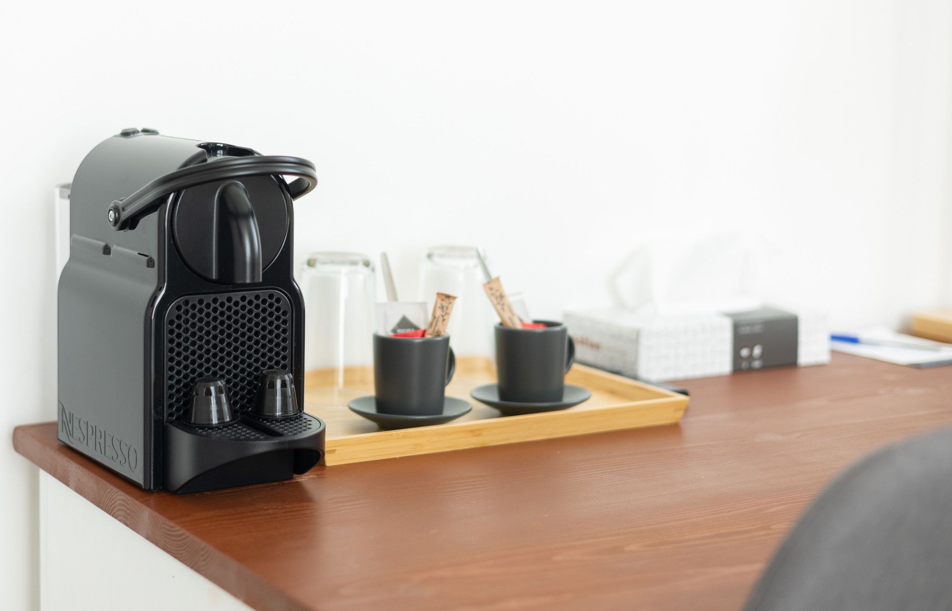 Black espresso machine, coffee cups, and accessories on a wooden counter against a white wall.