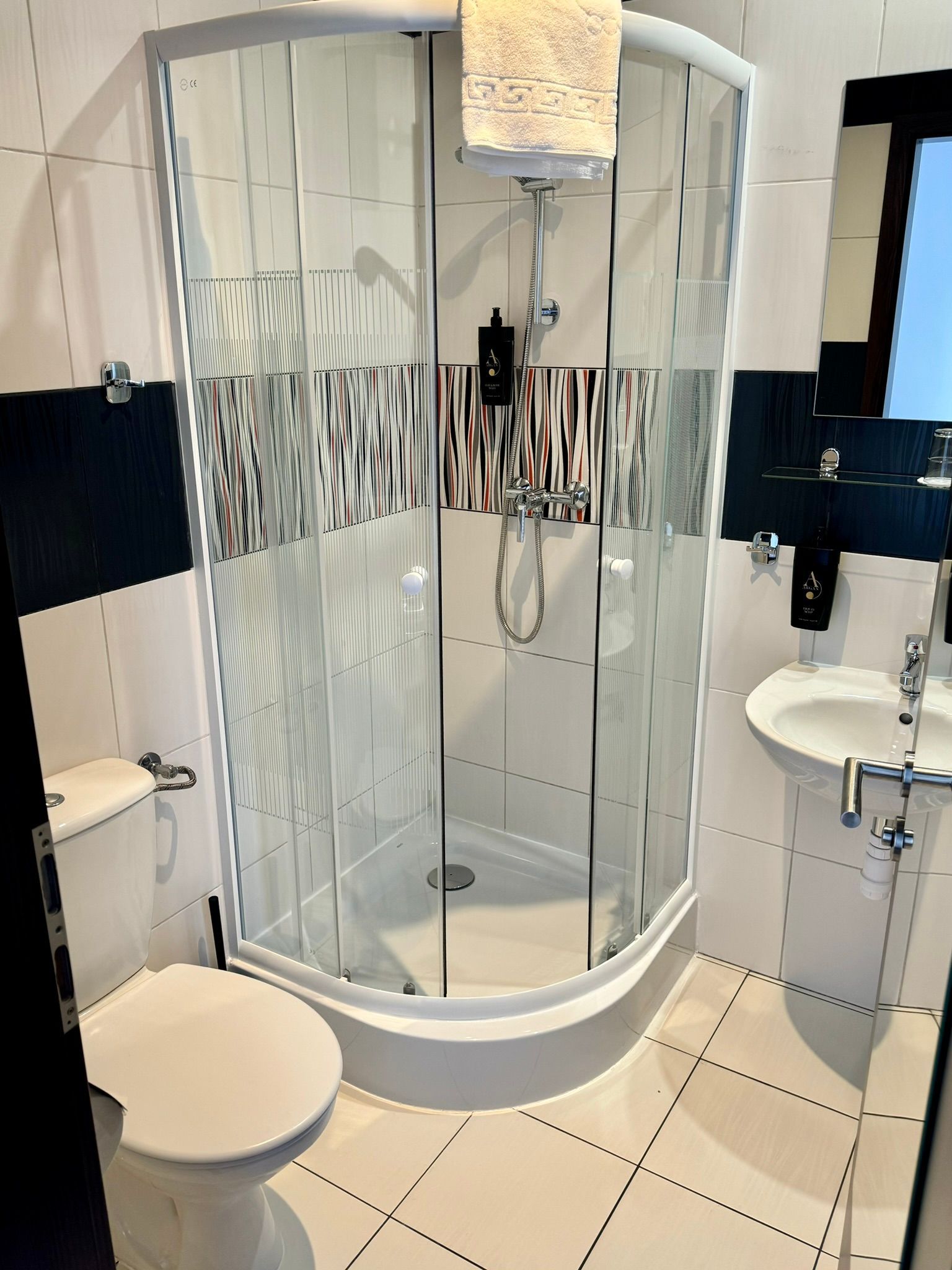 Small bathroom with curved shower, toilet, and sink. White, black, and silver tiles.