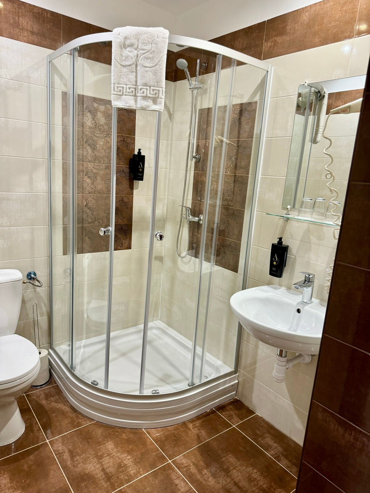 A small hotel bathroom with a glass shower, toilet, and sink. Brown and beige tiles.