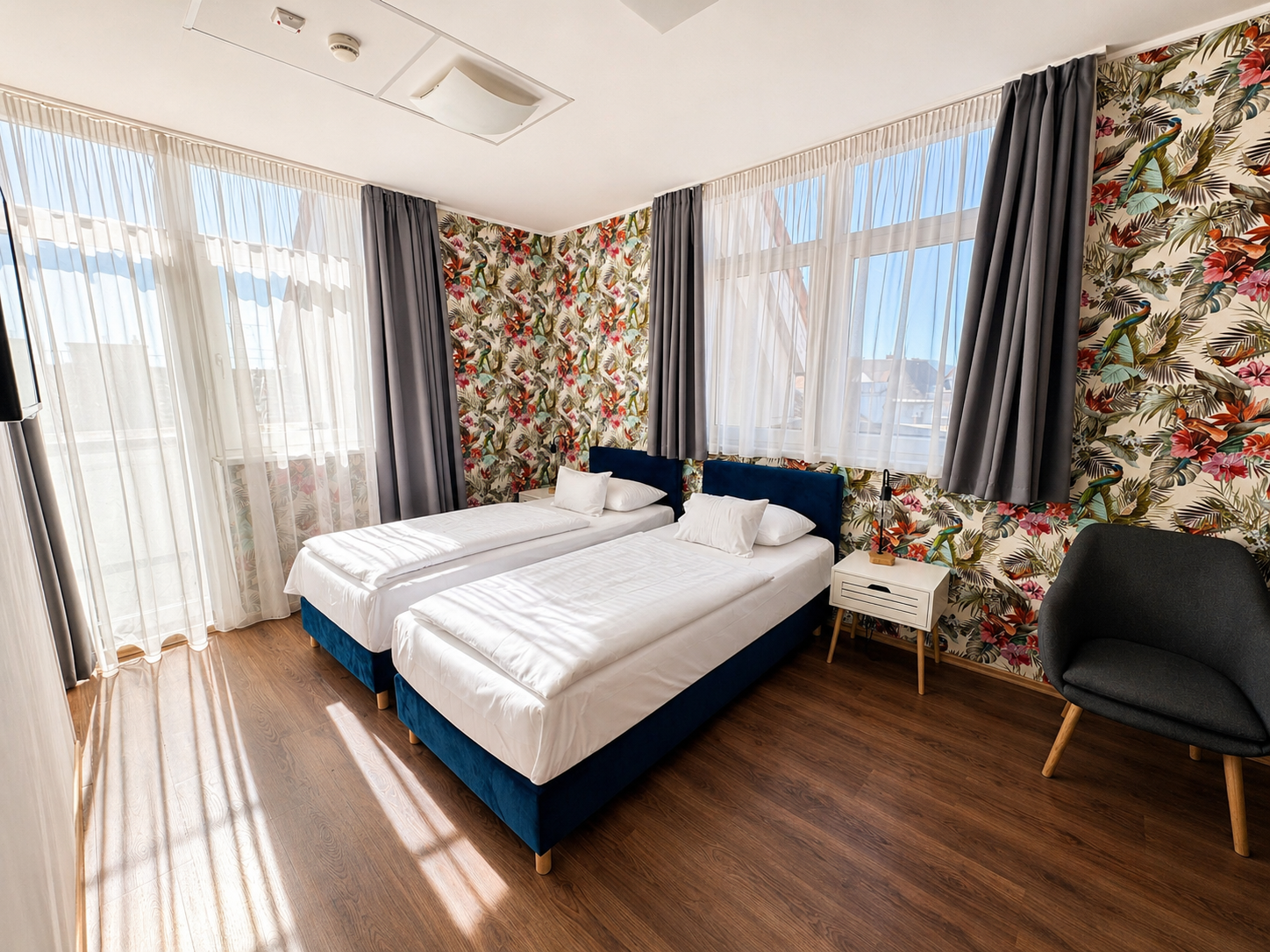 Hotel room with a bed, desk, chair, and large windows with curtains; natural light.
