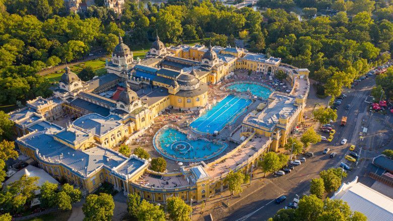 Aerial view of Széchenyi Thermal Baths in Budapest, Hungary; yellow building with blue pools, surrounded by green trees.