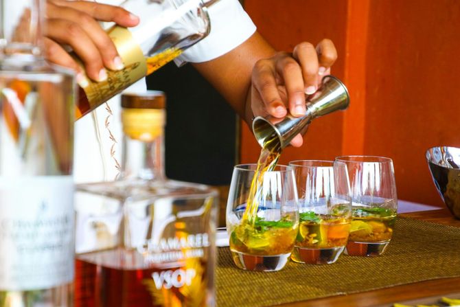 Bartender pouring amber liquid into glasses with mint, bottles in the foreground, orange background.