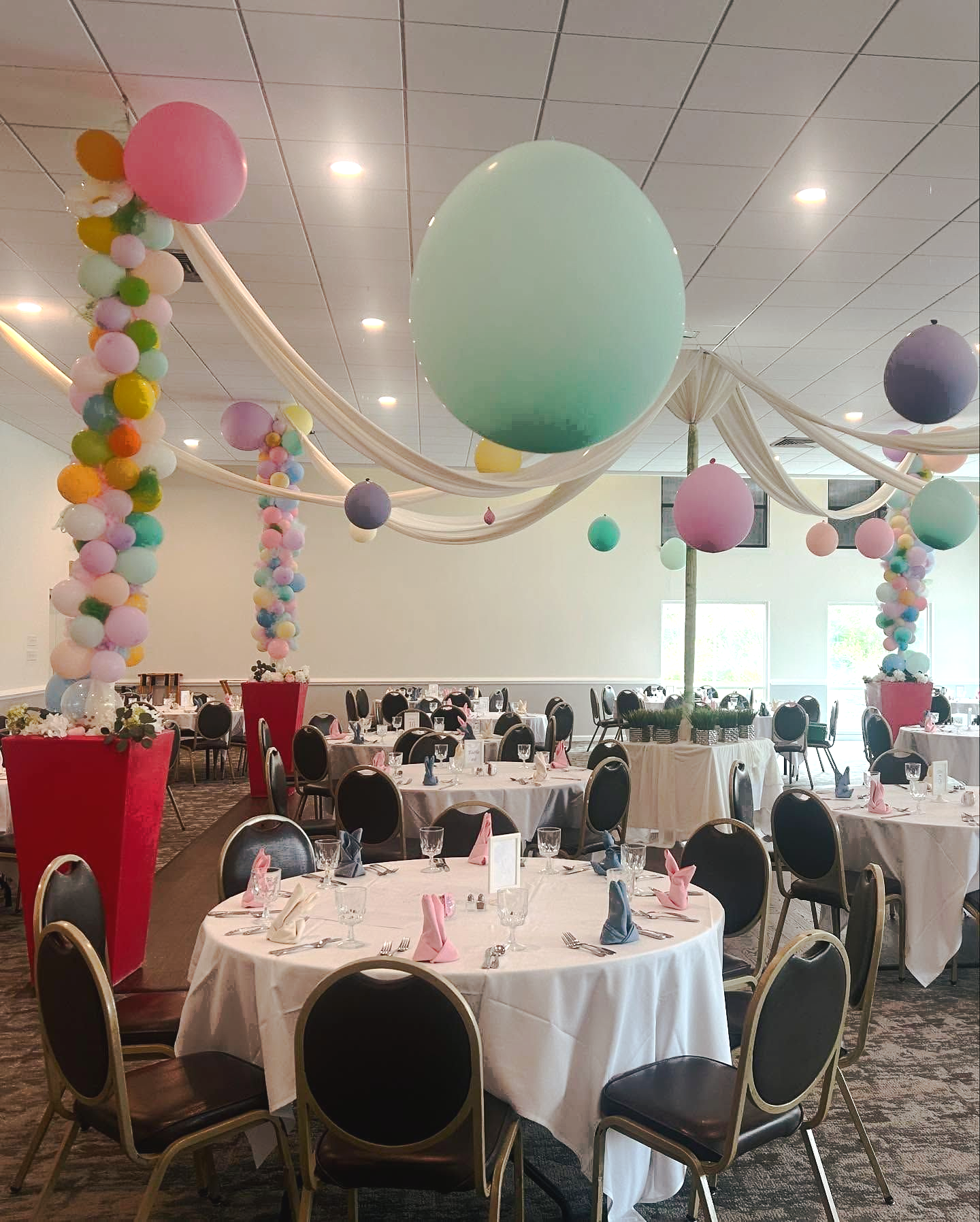 Balloon-decorated banquet hall with round tables set for an event and pastel hanging decorations.