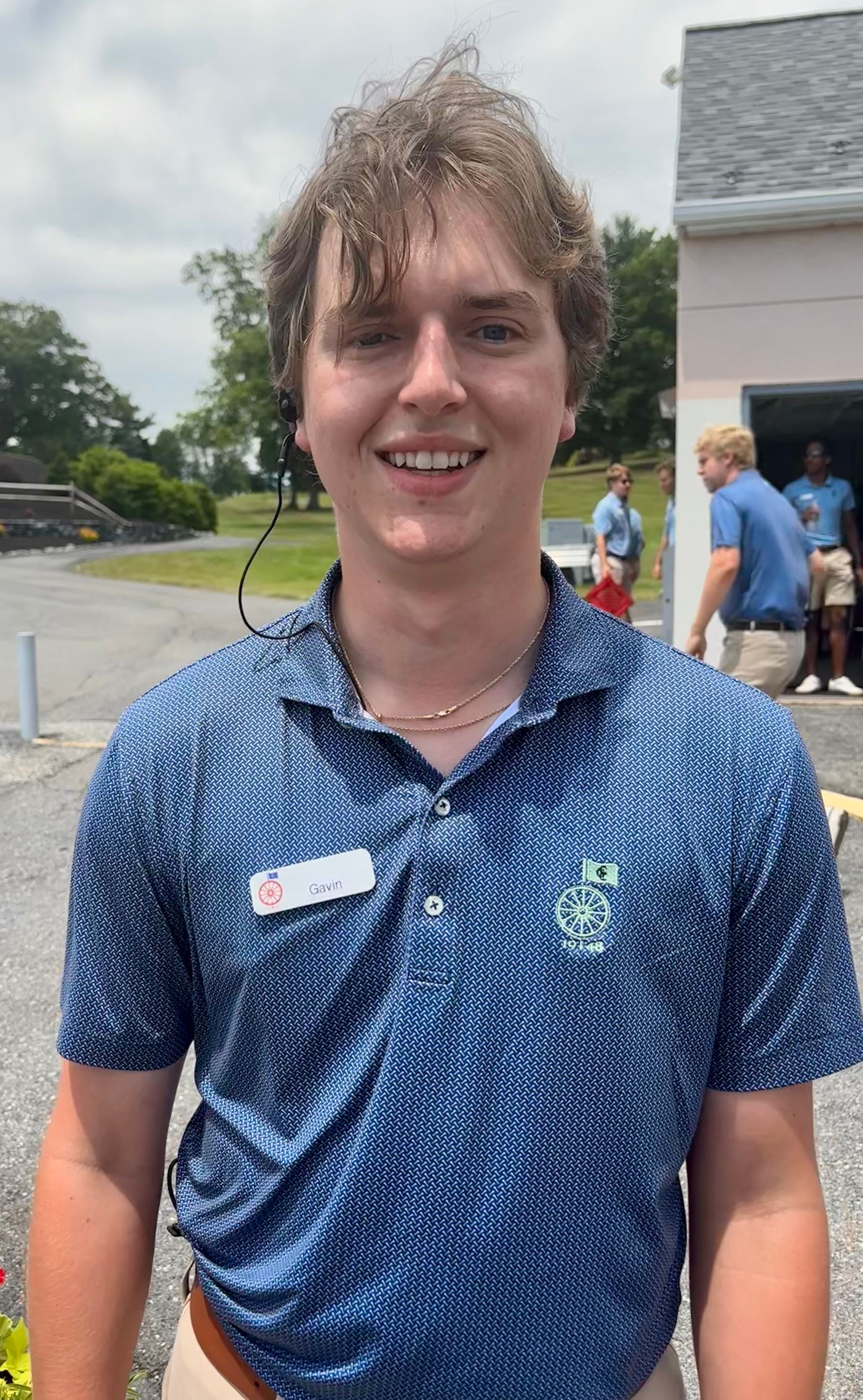 Smiling person in a blue patterned shirt standing outdoors near a building