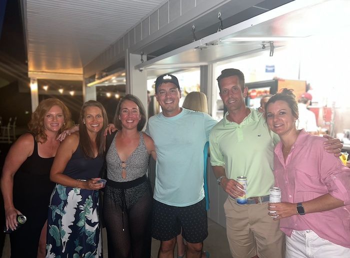 Six people smiling and posing with drinks on a covered outdoor patio at night.