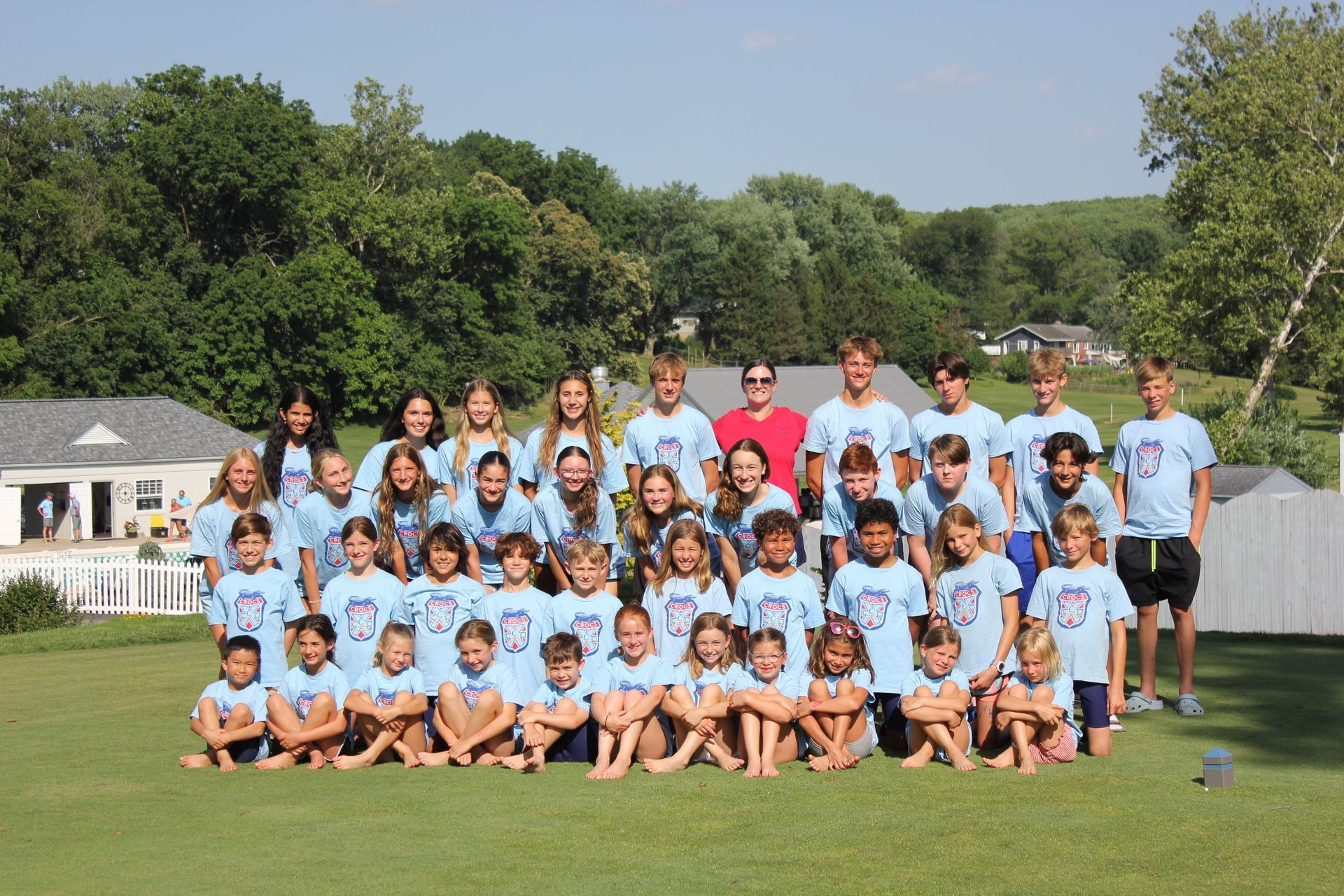 Group of people in light blue shirts posing on a grassy field with trees and houses in the background