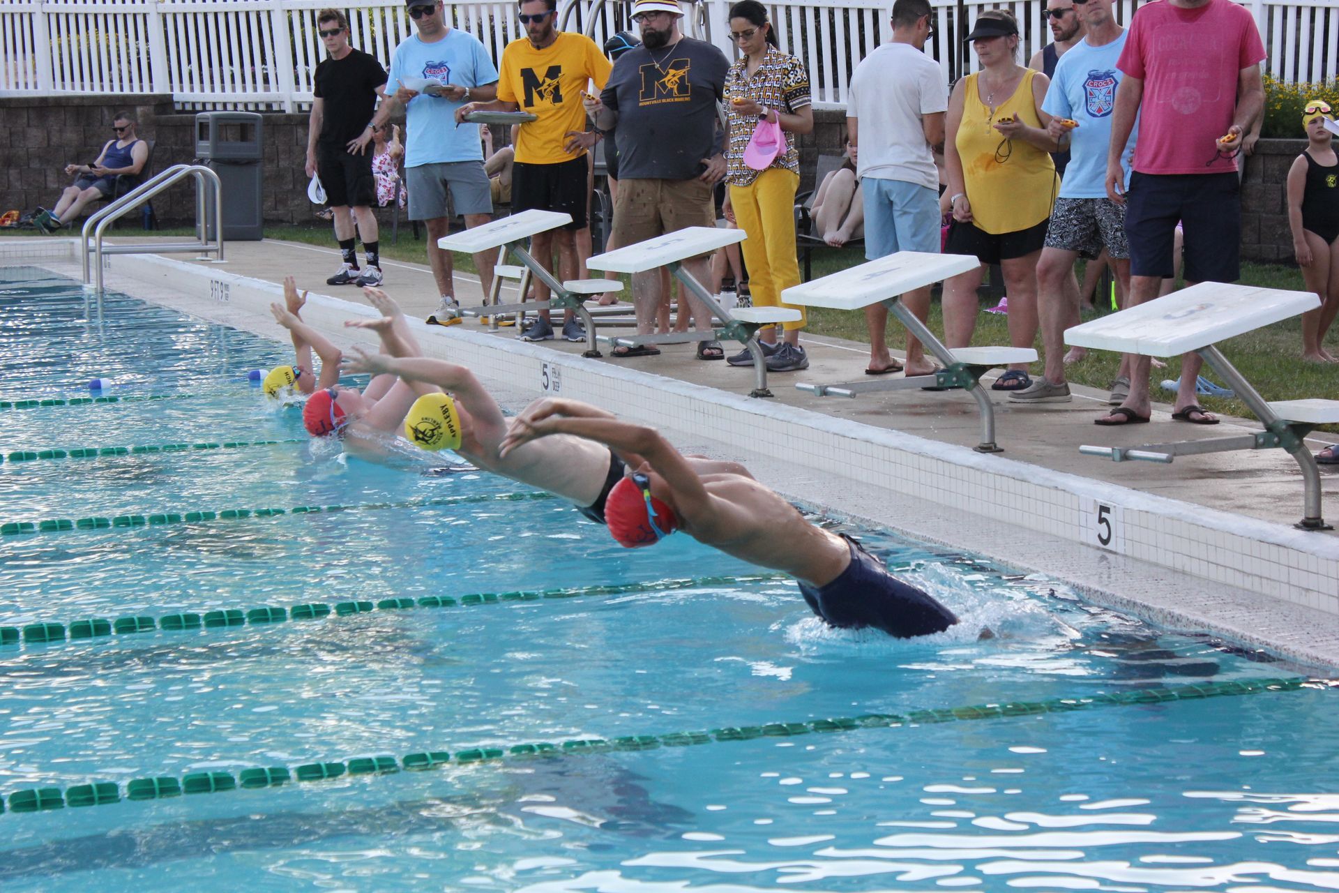 Swimmers diving into a pool at a race start, with spectators standing along the deck.