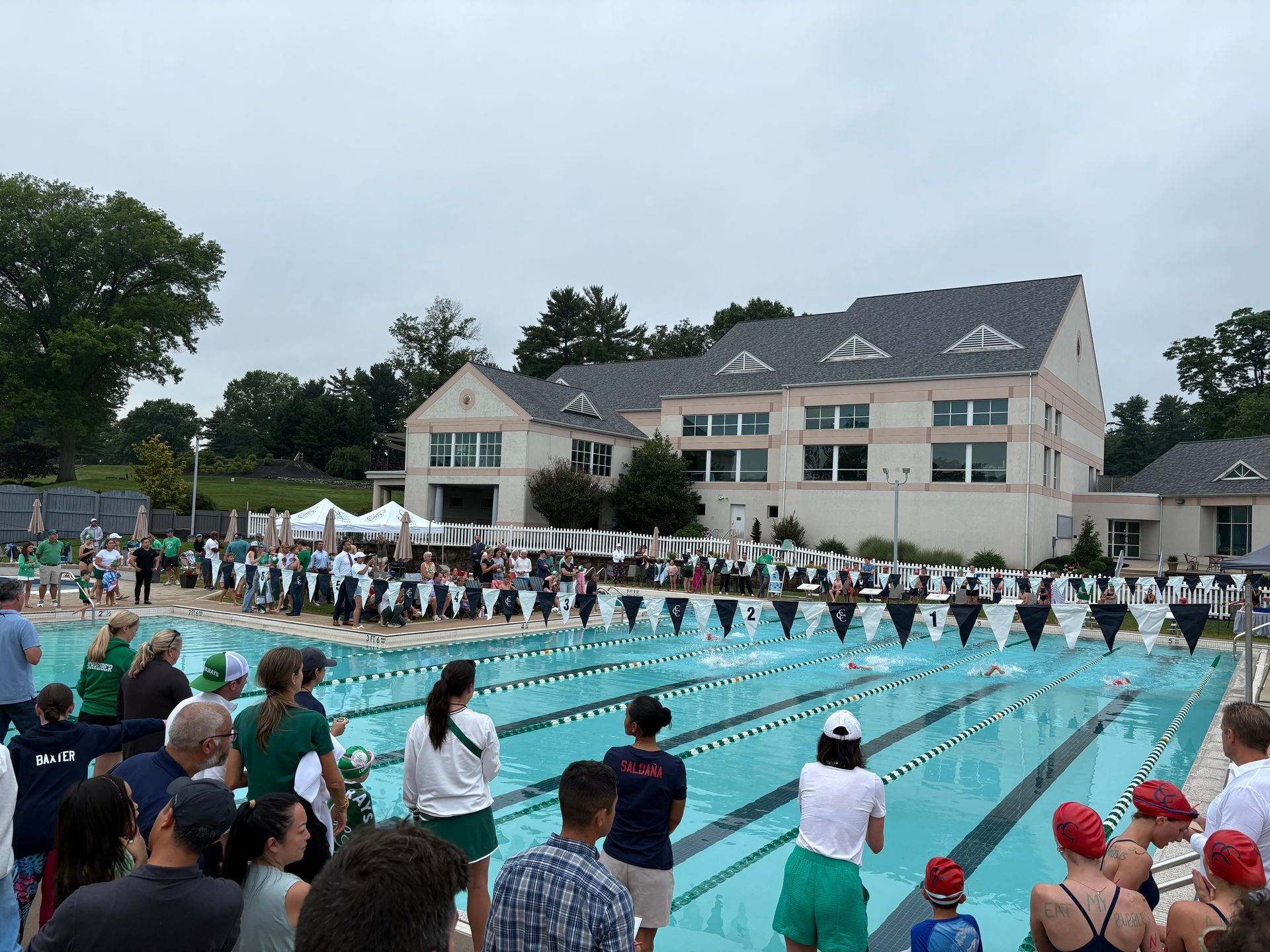 Crowded outdoor pool swim meet with lanes, swimmers on deck, and spectators by a large clubhouse