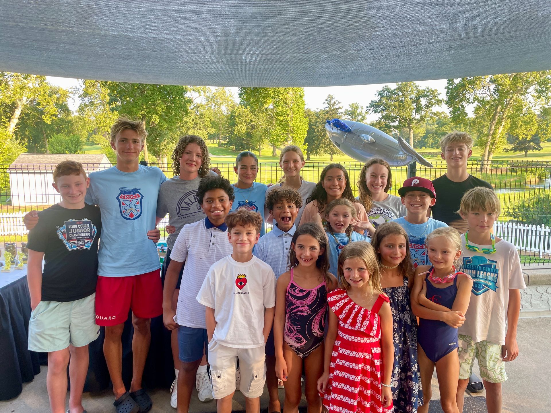 Group of smiling children standing together outdoors under a canopy on a sunny day.