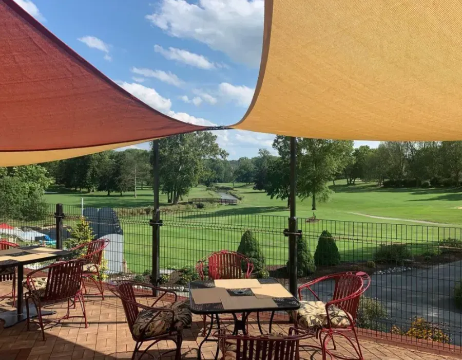 Patio tables under orange shade sails overlook a green park and fountain on a sunny day