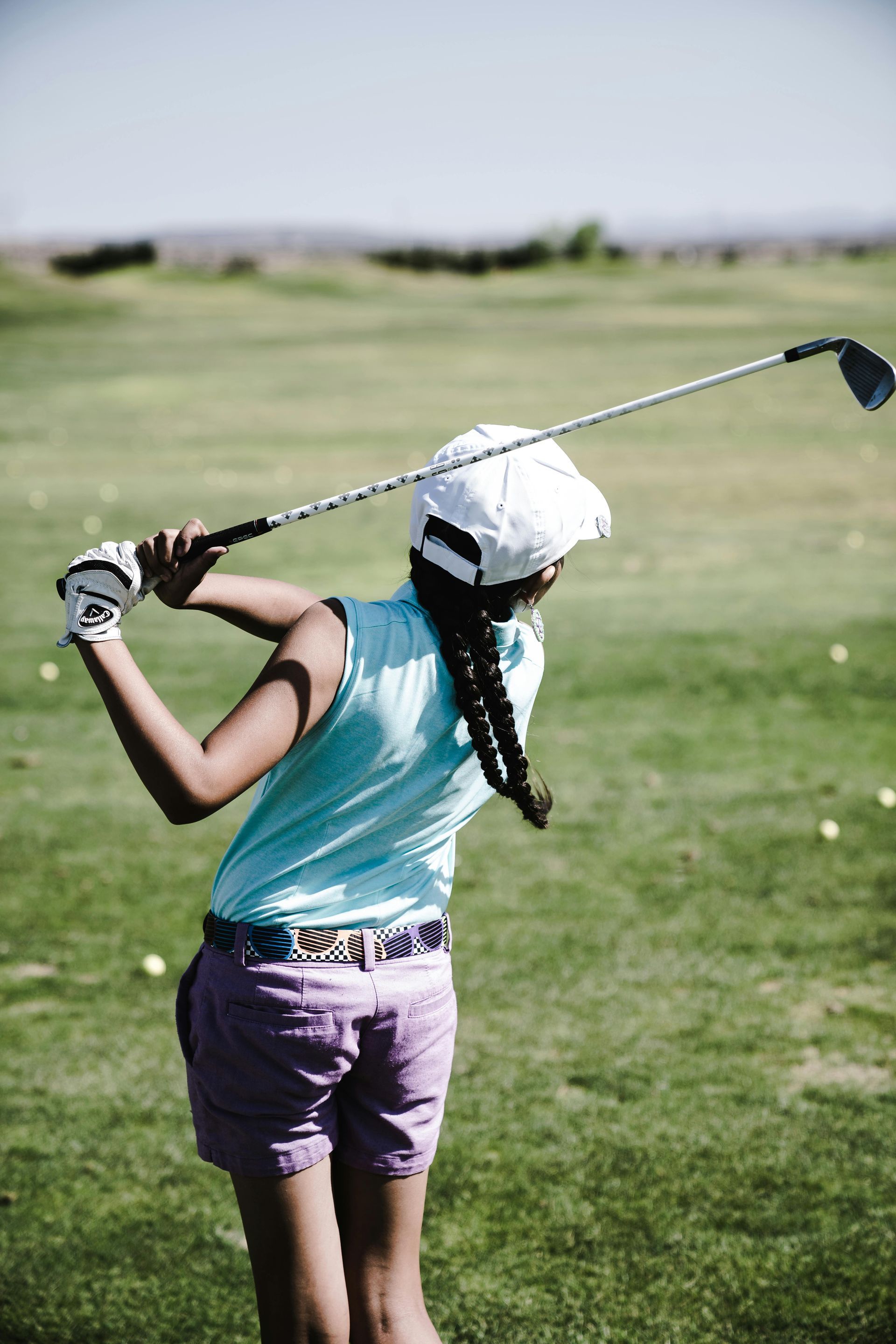 Golfer in teal shirt and purple shorts swings a club on a grassy course