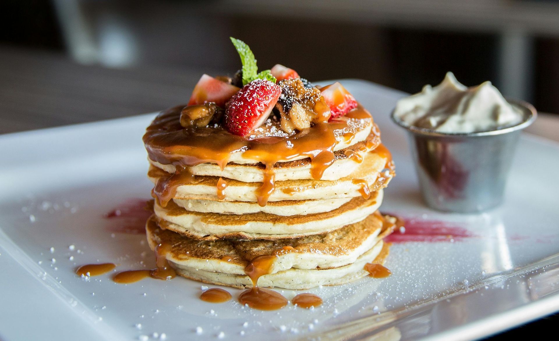 Stack of pancakes topped with strawberries and syrup on a white plate, with a side of whipped cream