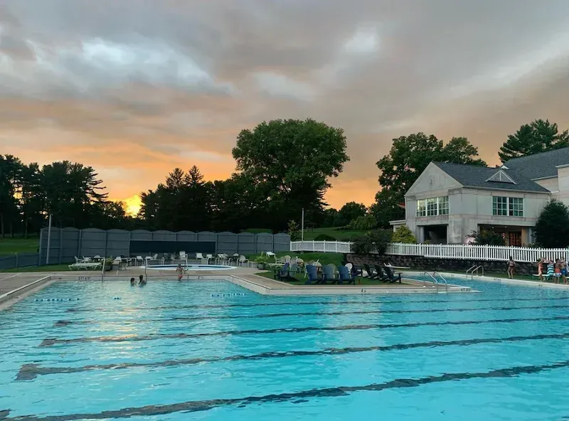 Outdoor swimming pool at sunset with swimmers, lounge chairs, and a house in the background