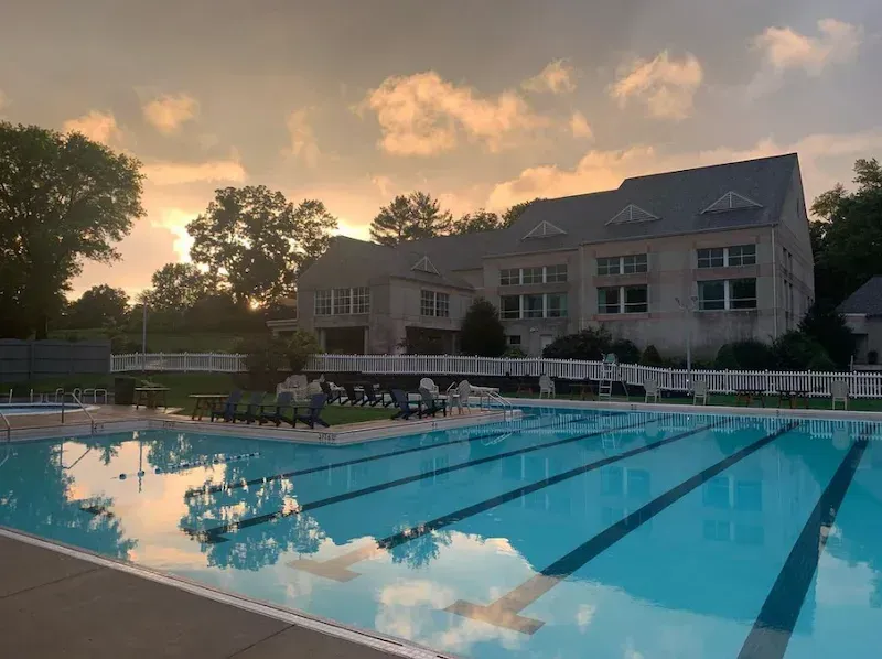 Outdoor swimming pool at sunset beside a clubhouse, with lounge chairs and trees reflecting in the water