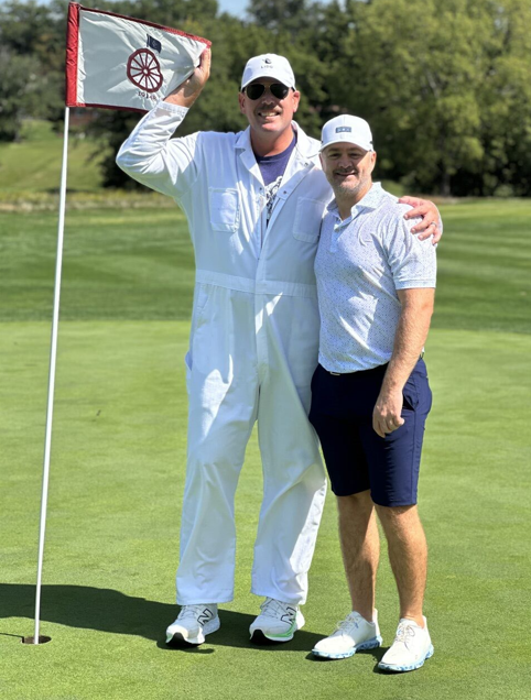 Two people posing on a golf green beside a flag, one holding the flag and both smiling.