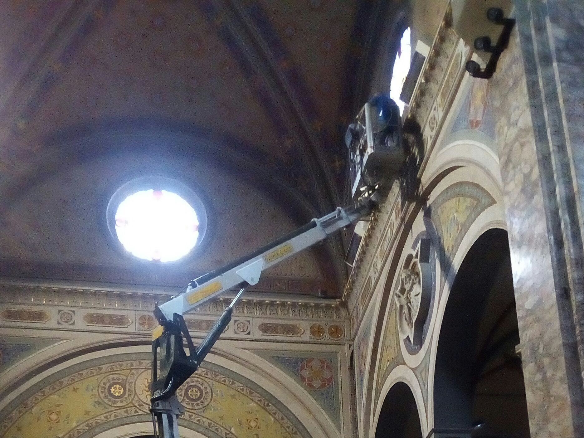 Una piattaforma elevatrice si estende verso l'interno decorato della chiesa, vicino a una finestra circolare. È visibile un operaio nell'ascensore.
