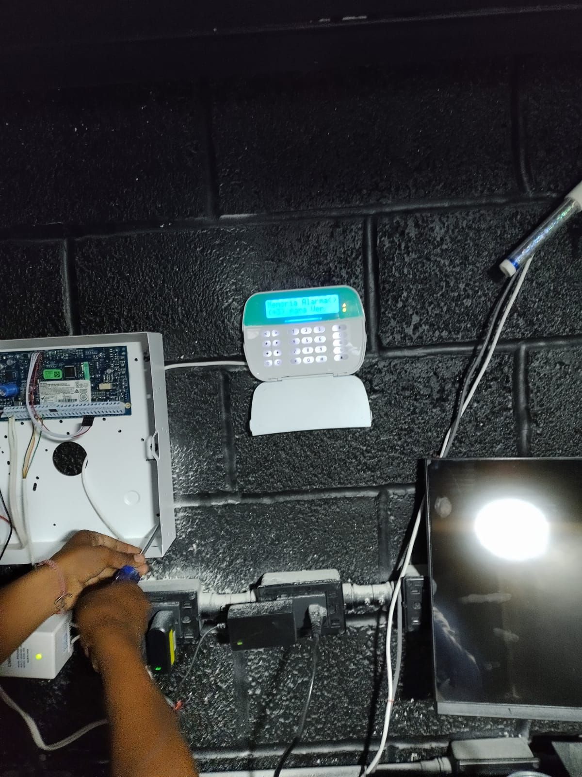 Una persona trabaja en un panel de seguridad sobre una pared de ladrillos negros. Hay un monitor a la derecha del panel.