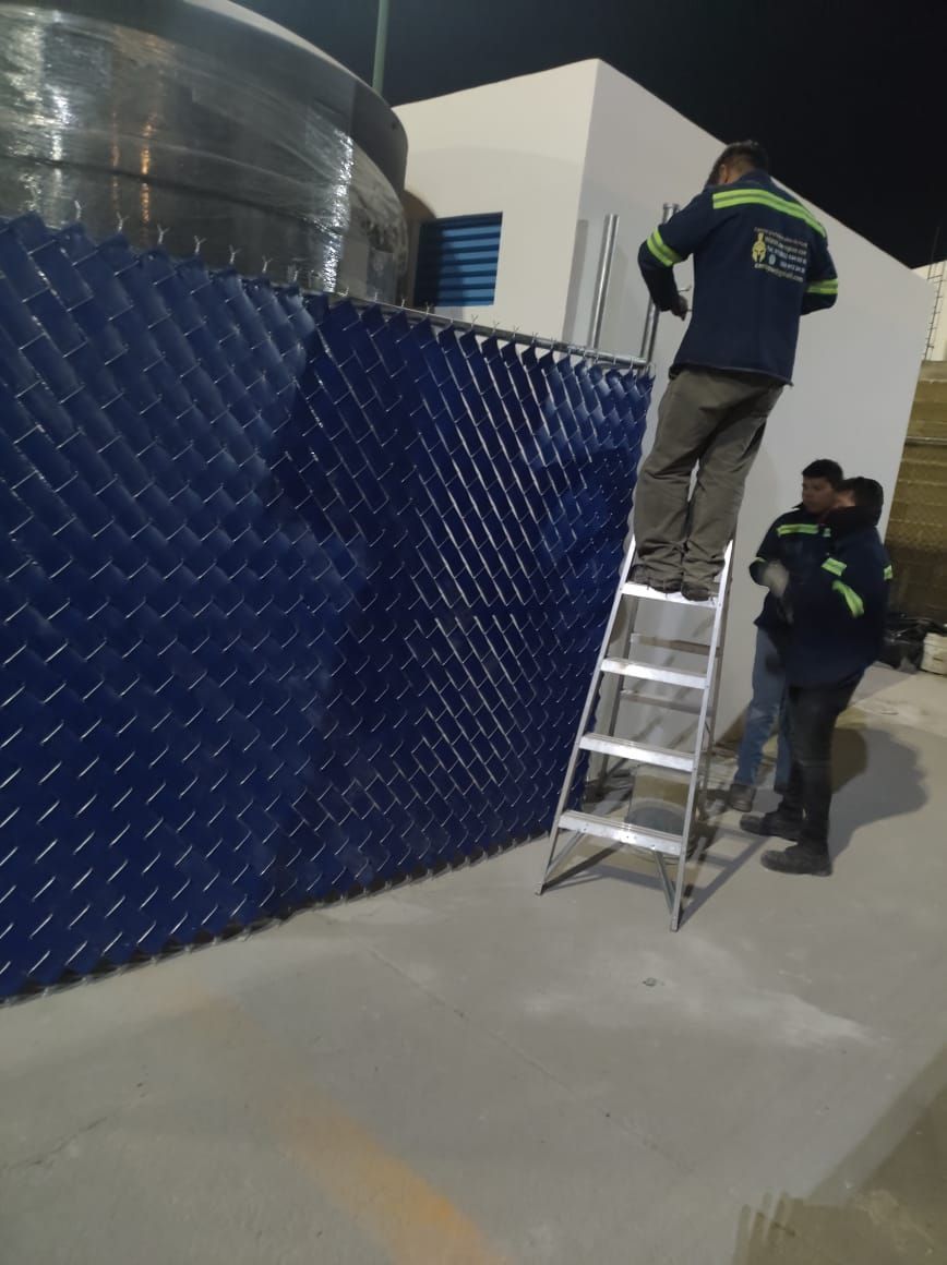 Trabajadores en una escalera instalando un panel de plástico azul cerca de un tanque de agua en un sitio industrial.