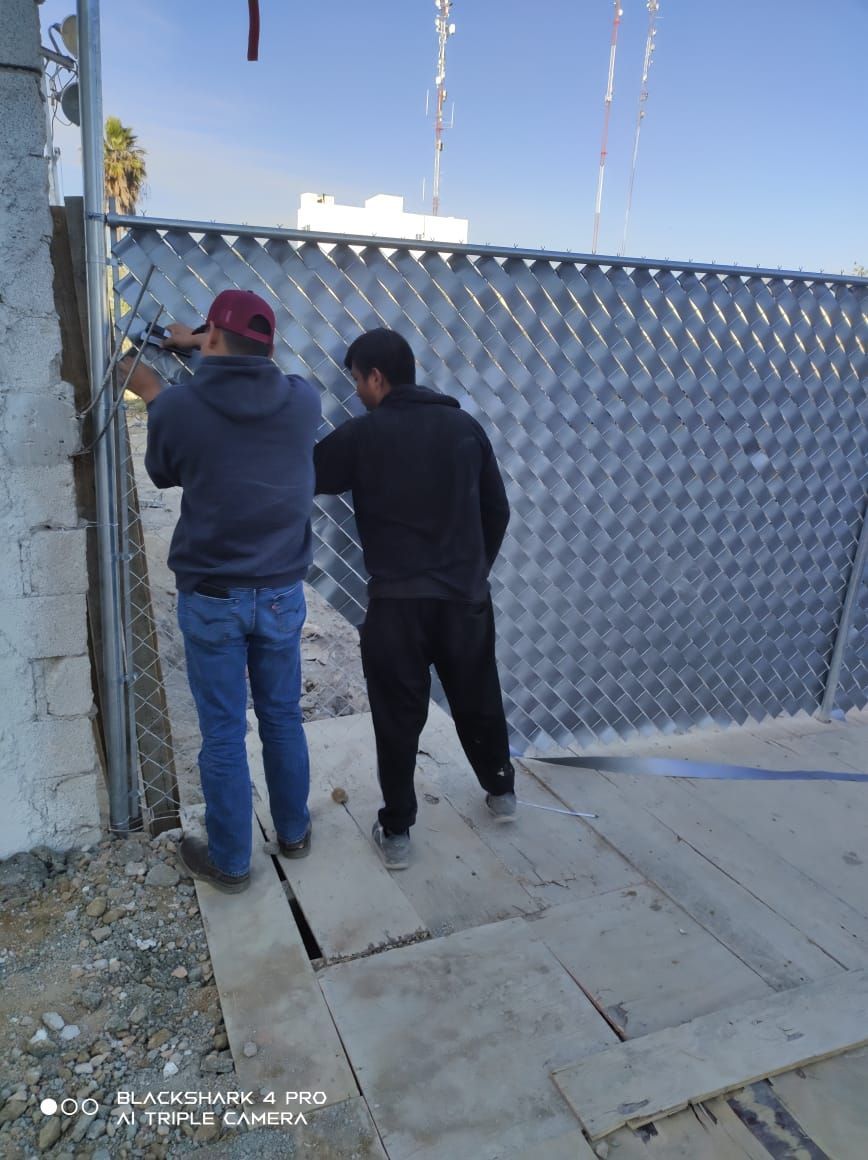 Dos hombres trabajando en una puerta metálica al aire libre. Uno sostiene unos alicates, el otro observa. Día soleado.