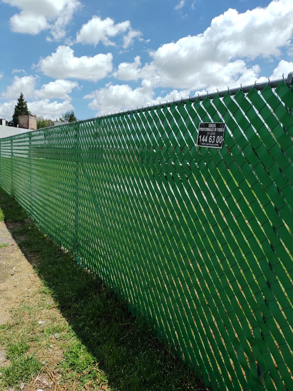 Valla de alambre verde con listones para privacidad, junto a una zona de césped, bajo un cielo parcialmente nublado.
