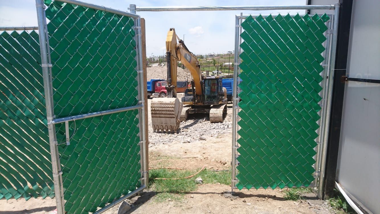 La puerta de alambre verde de la cerca se abre y revela una excavadora en un sitio de construcción.
