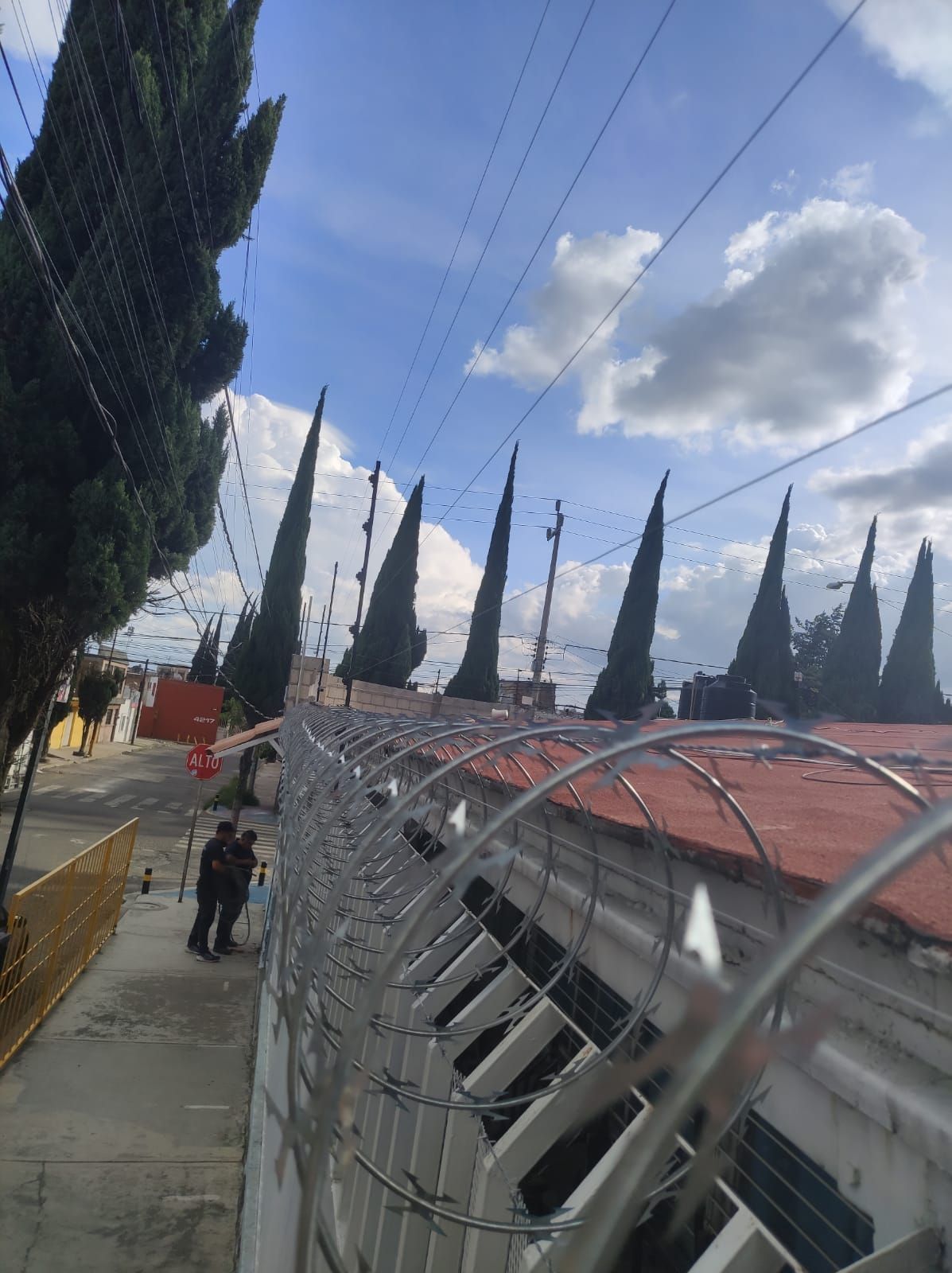Muro coronado con alambre de púas, árboles altos al fondo, dos personas caminando por una acera, cielo azul con nubes.