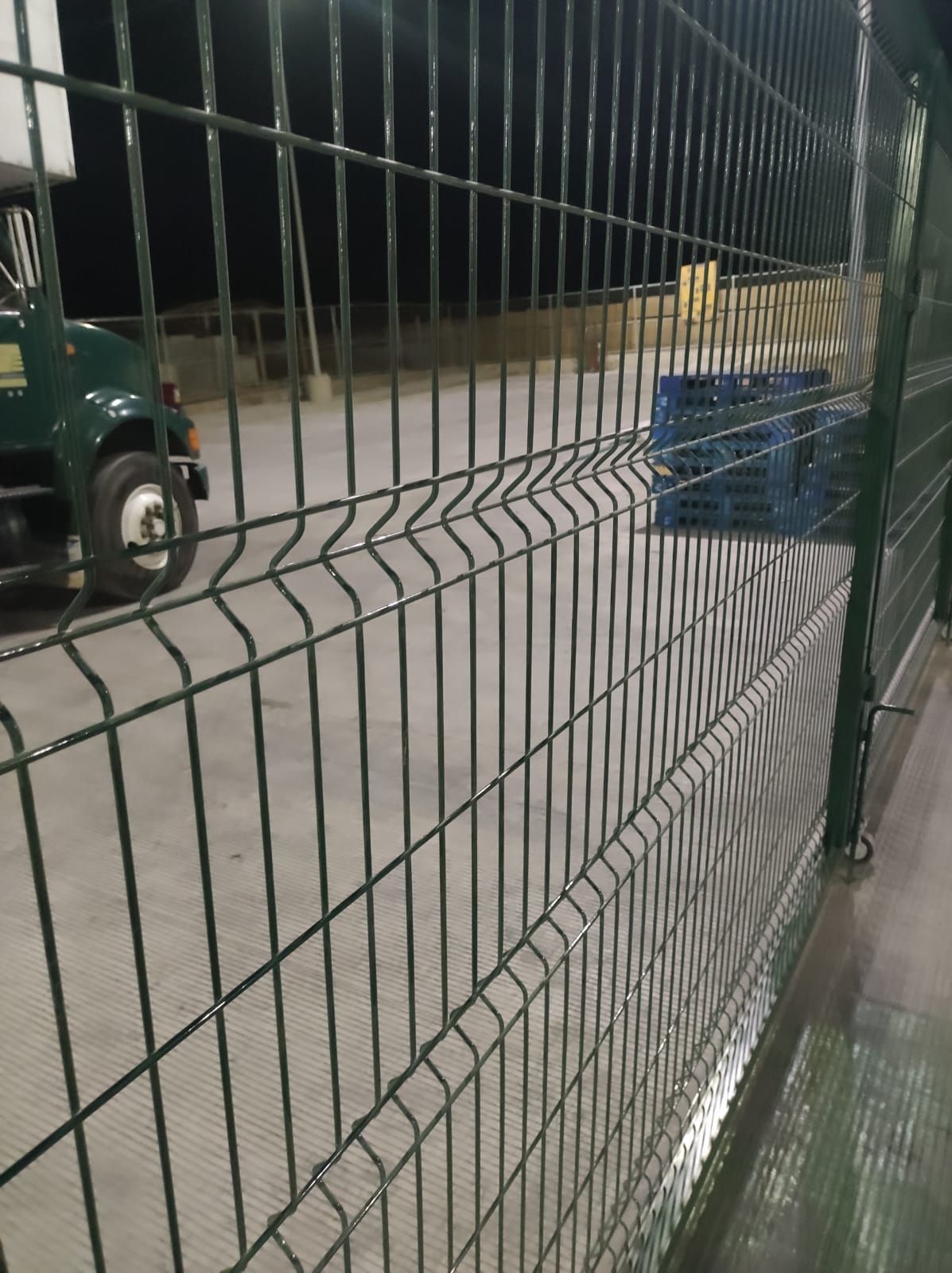 Valla de malla verde enfocada, con un vehículo verde y palés al fondo. Escena nocturna.