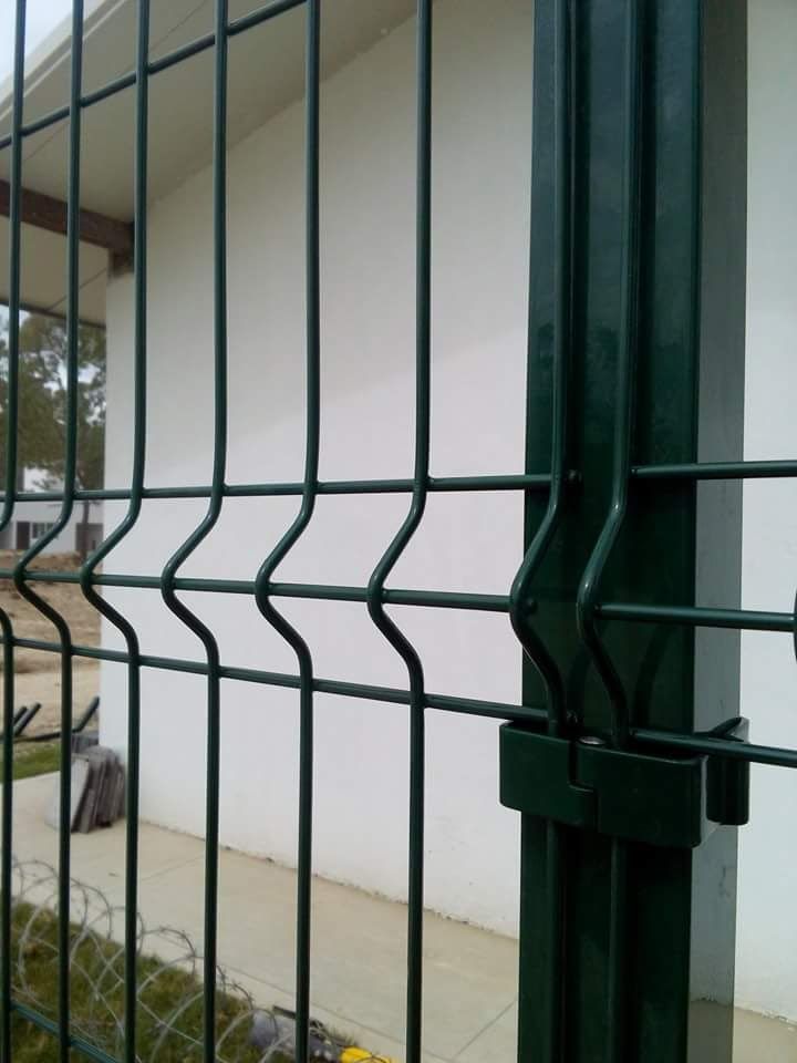 Valla metálica verde con alambres verticales y horizontales; fijada a un poste verde oscuro frente a un edificio blanco.