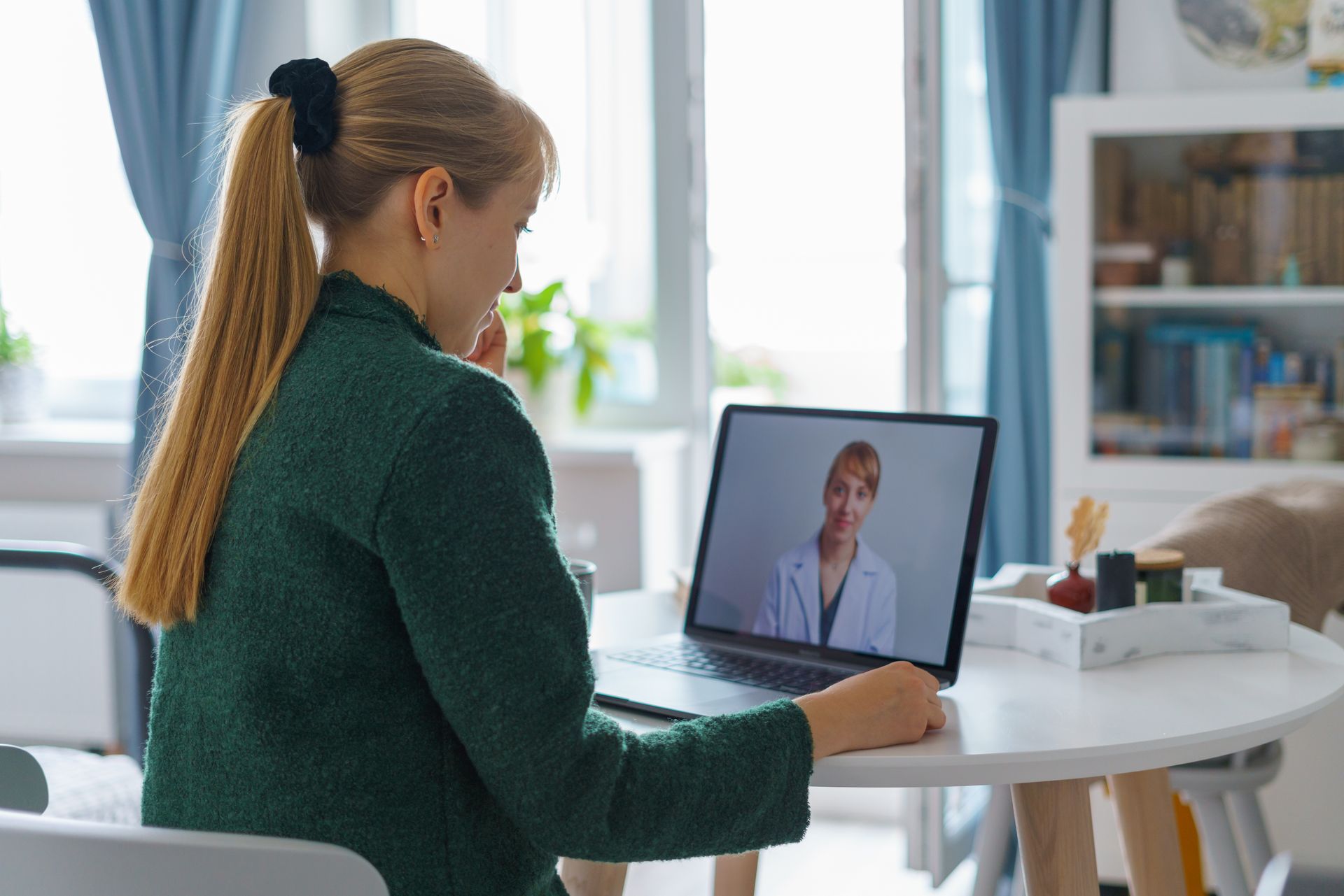 A woman is sitting at a table using a laptop computer to have a video call with a doctor.