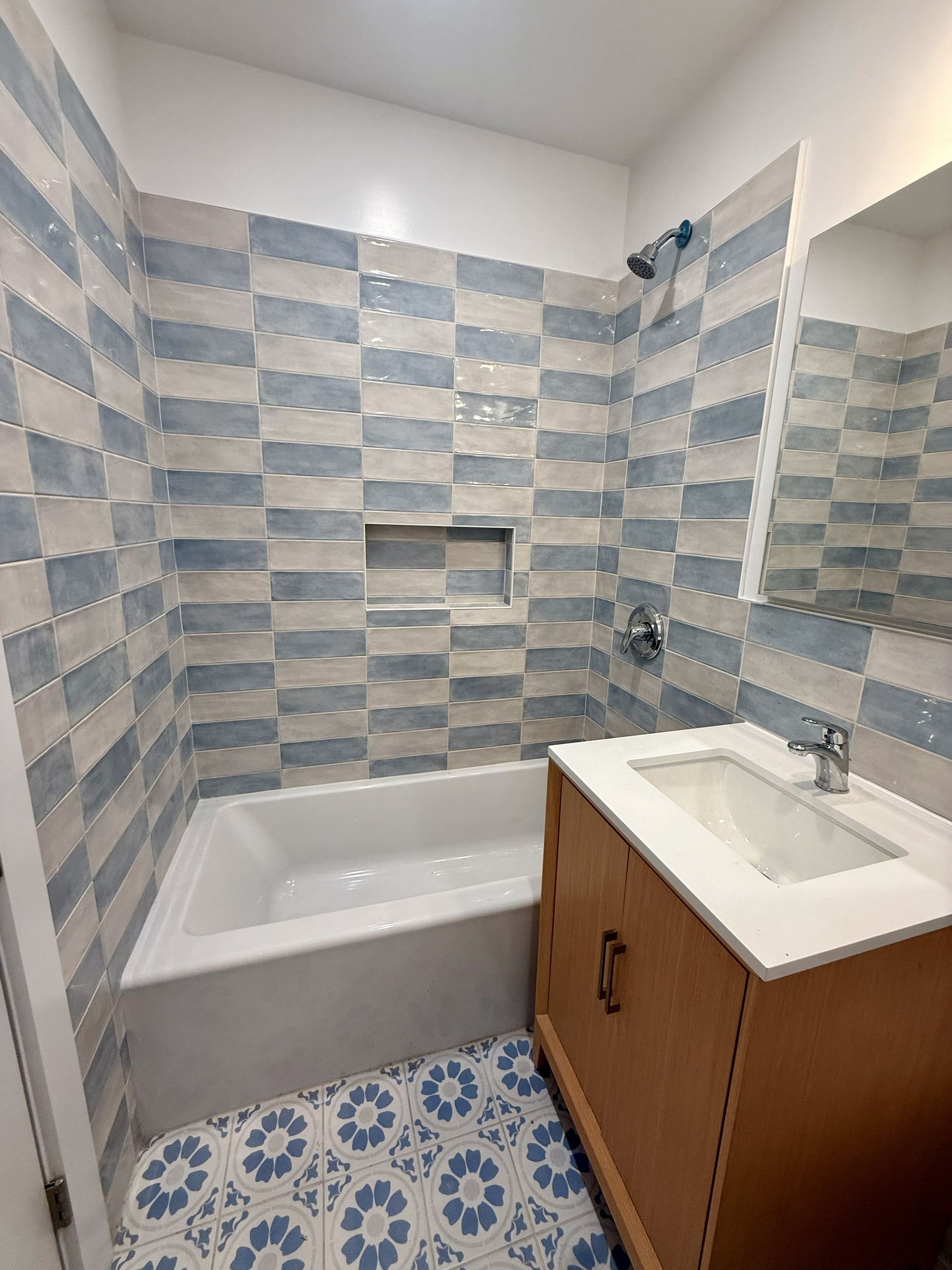 Small bathroom with a bathtub, sink vanity, blue-gray tiled walls, and patterned tile floor.