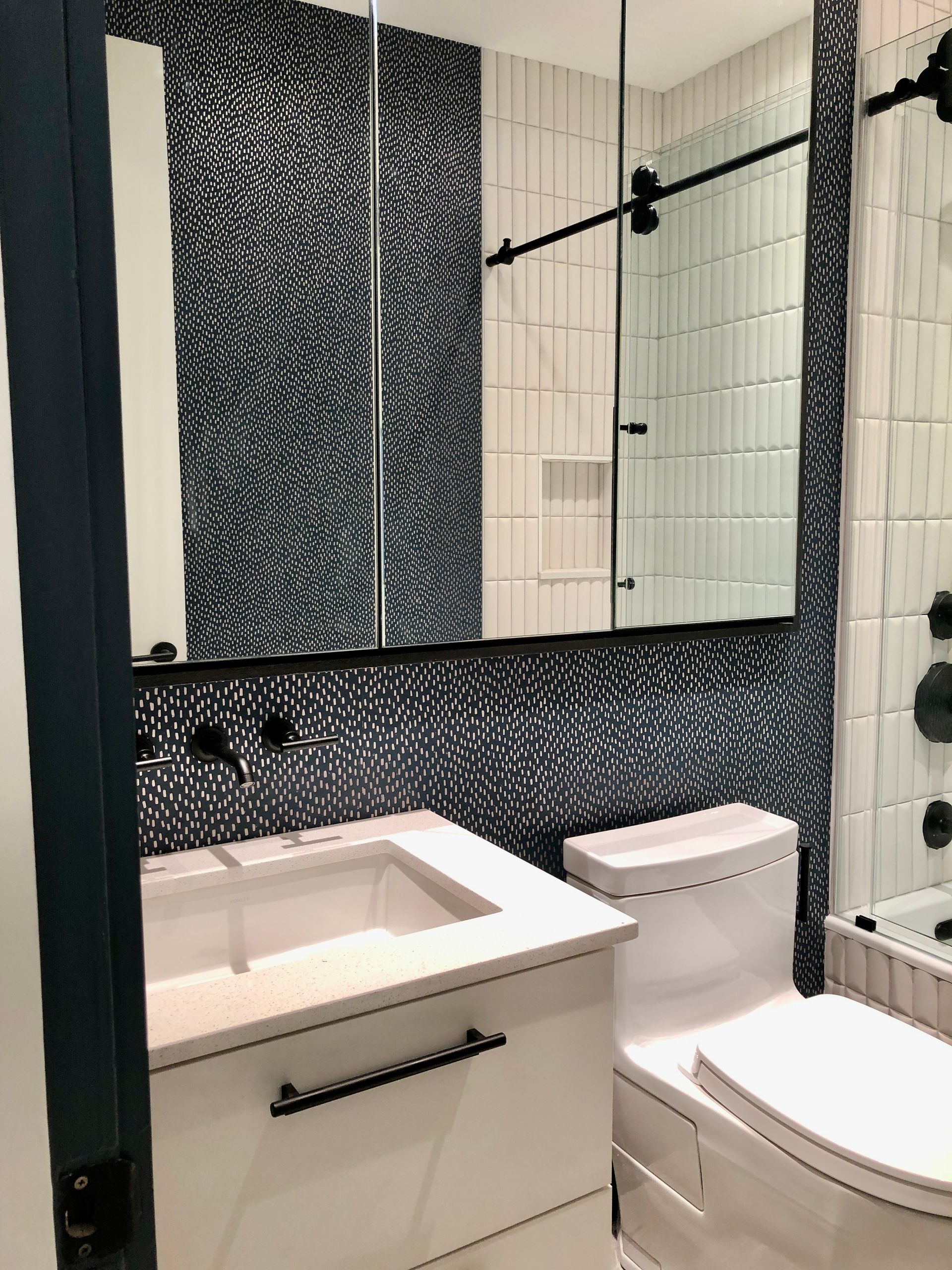 Small bathroom with white sink, toilet, black-speckled tile, mirror, and glass shower wall.