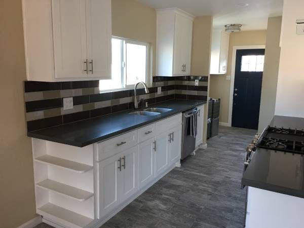 Narrow kitchen with white cabinets, black countertops, striped backsplash, and gray wood-look floor.