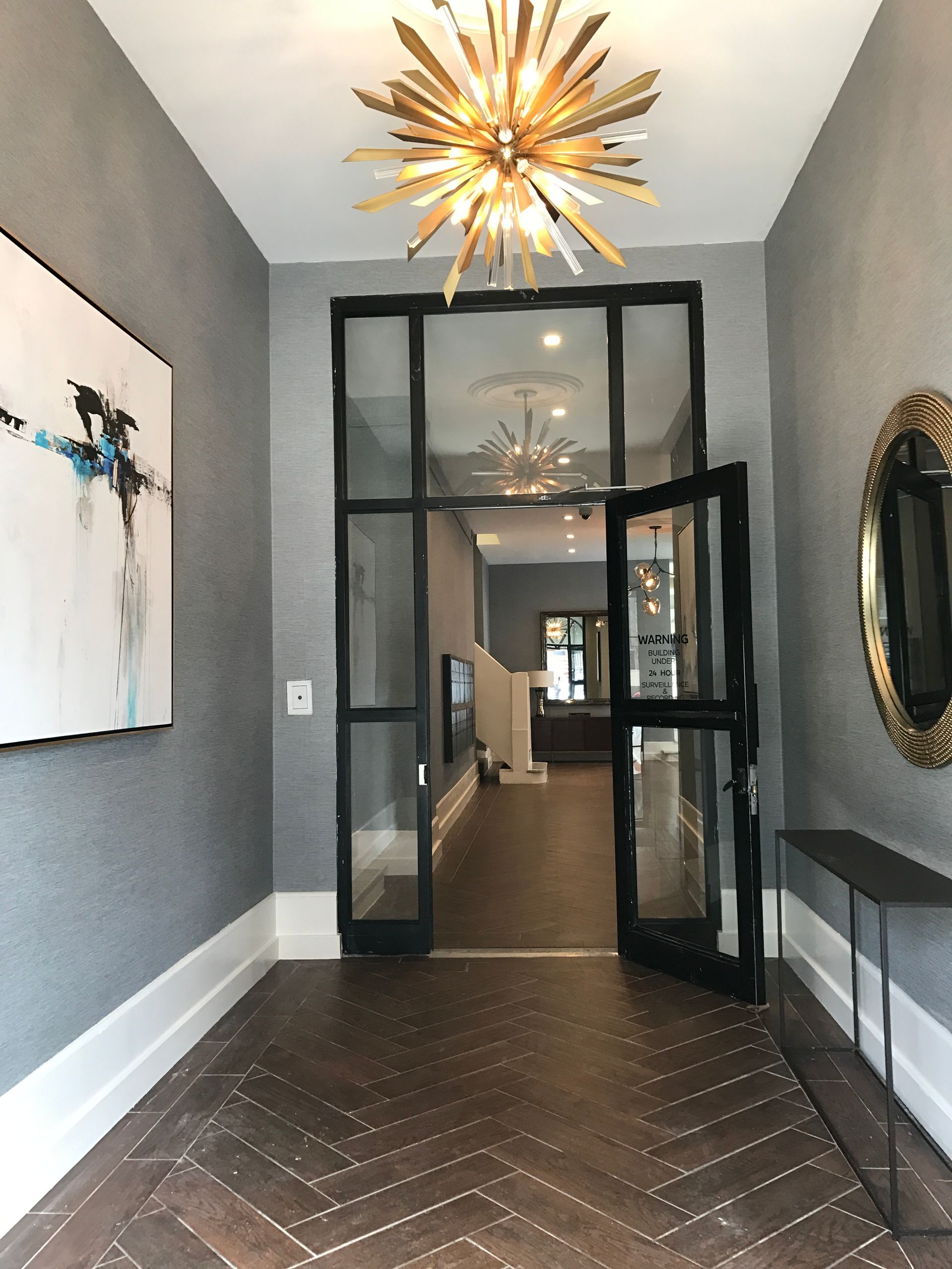 Modern hallway with gray walls, dark herringbone floor, black glass doors, and a gold starburst chandelier.