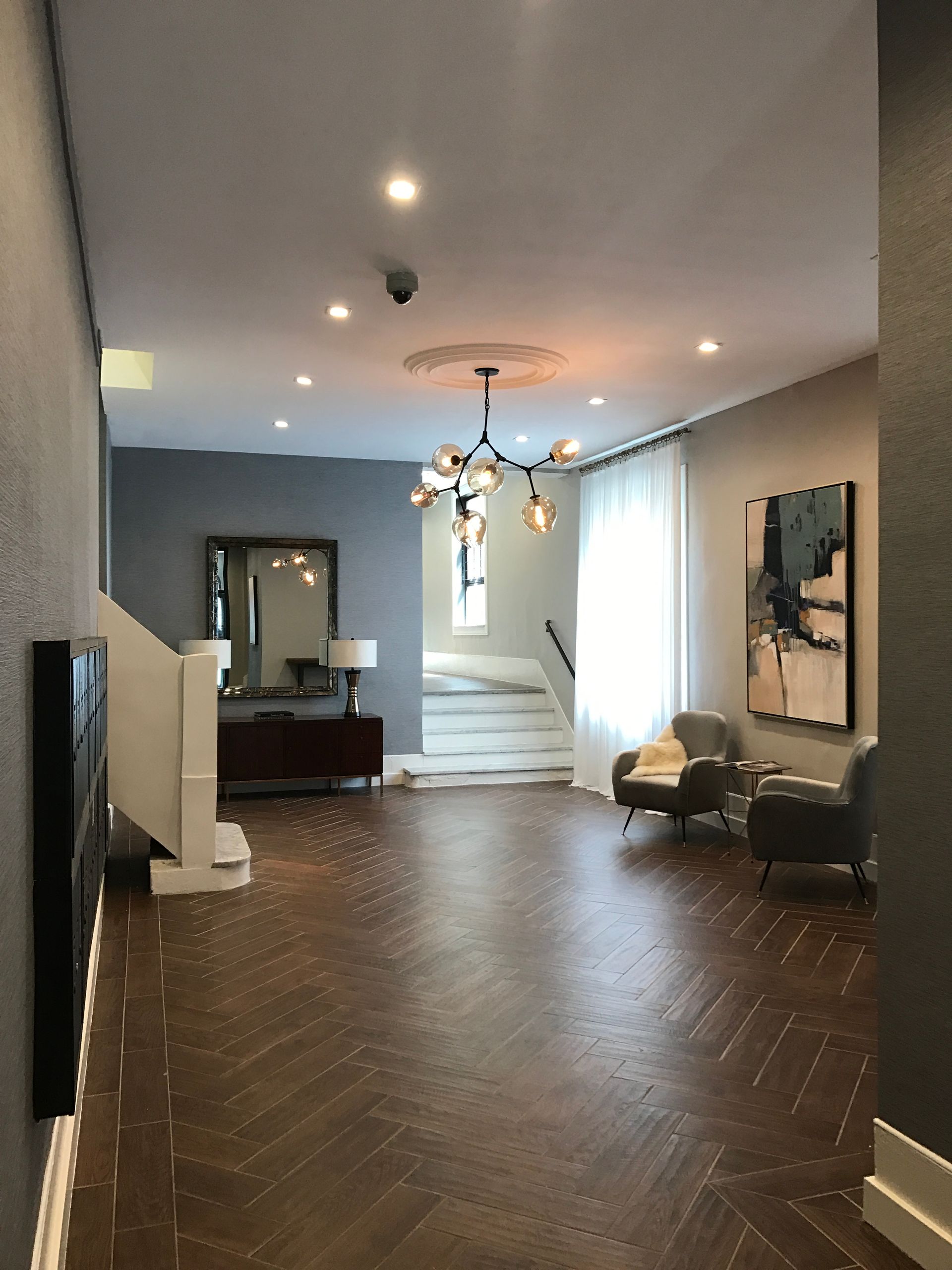 Modern living room with herringbone wood floor, chandelier, gray walls, and stairs leading to a bright hallway
