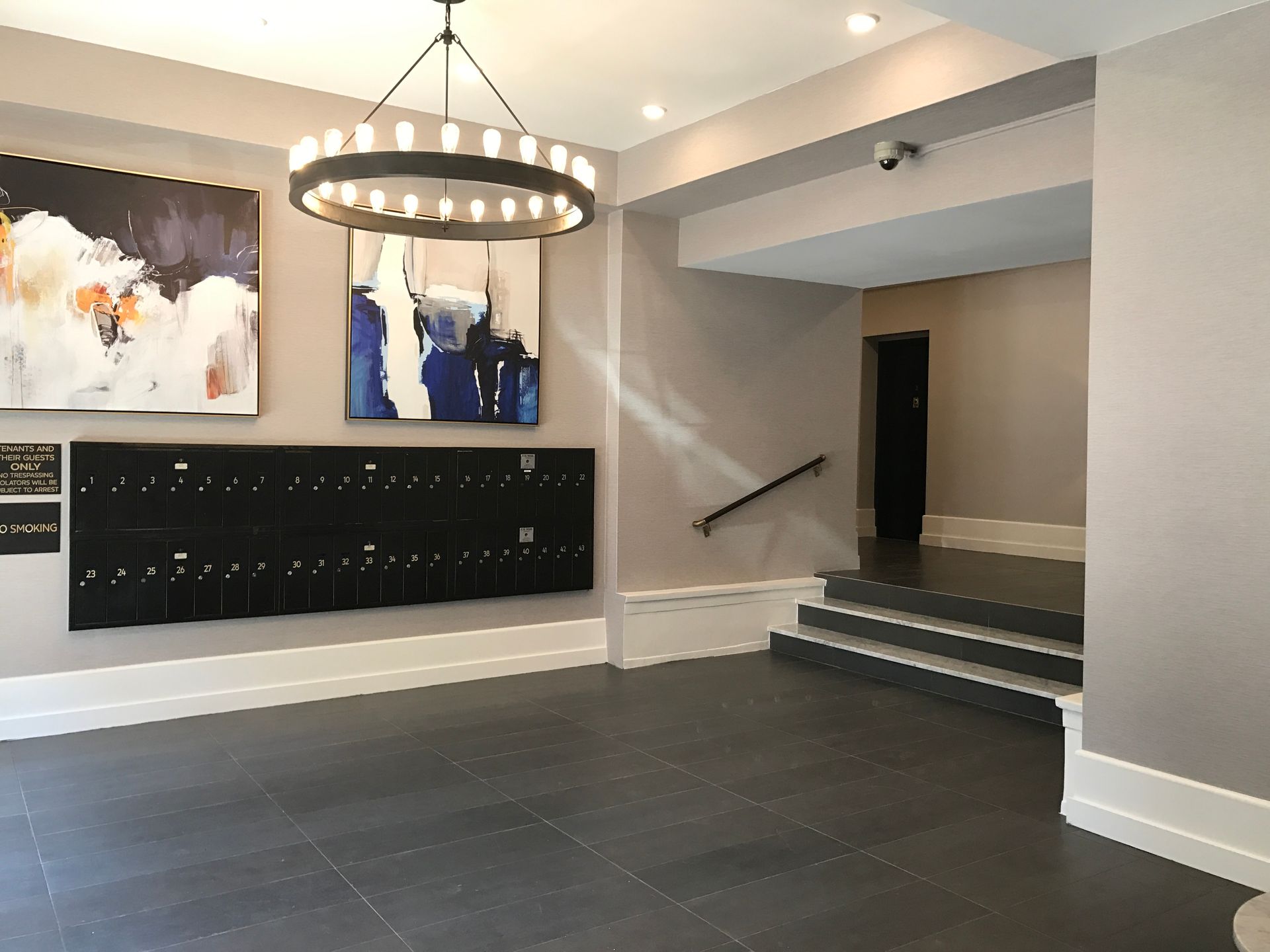 Modern lobby with dark tile floor, large chandelier, abstract art, and a staircase leading to a hallway