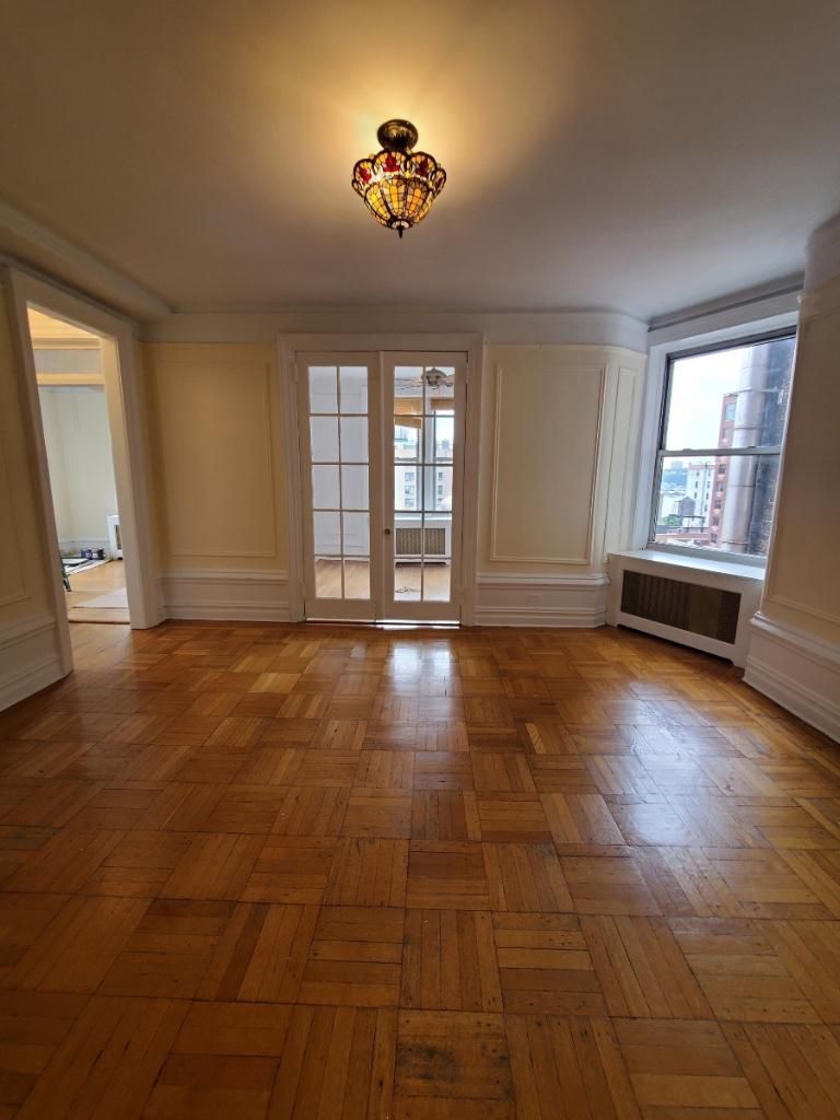 Empty living room with parquet wood floors, white walls, French doors, and large windows with radiator covers.