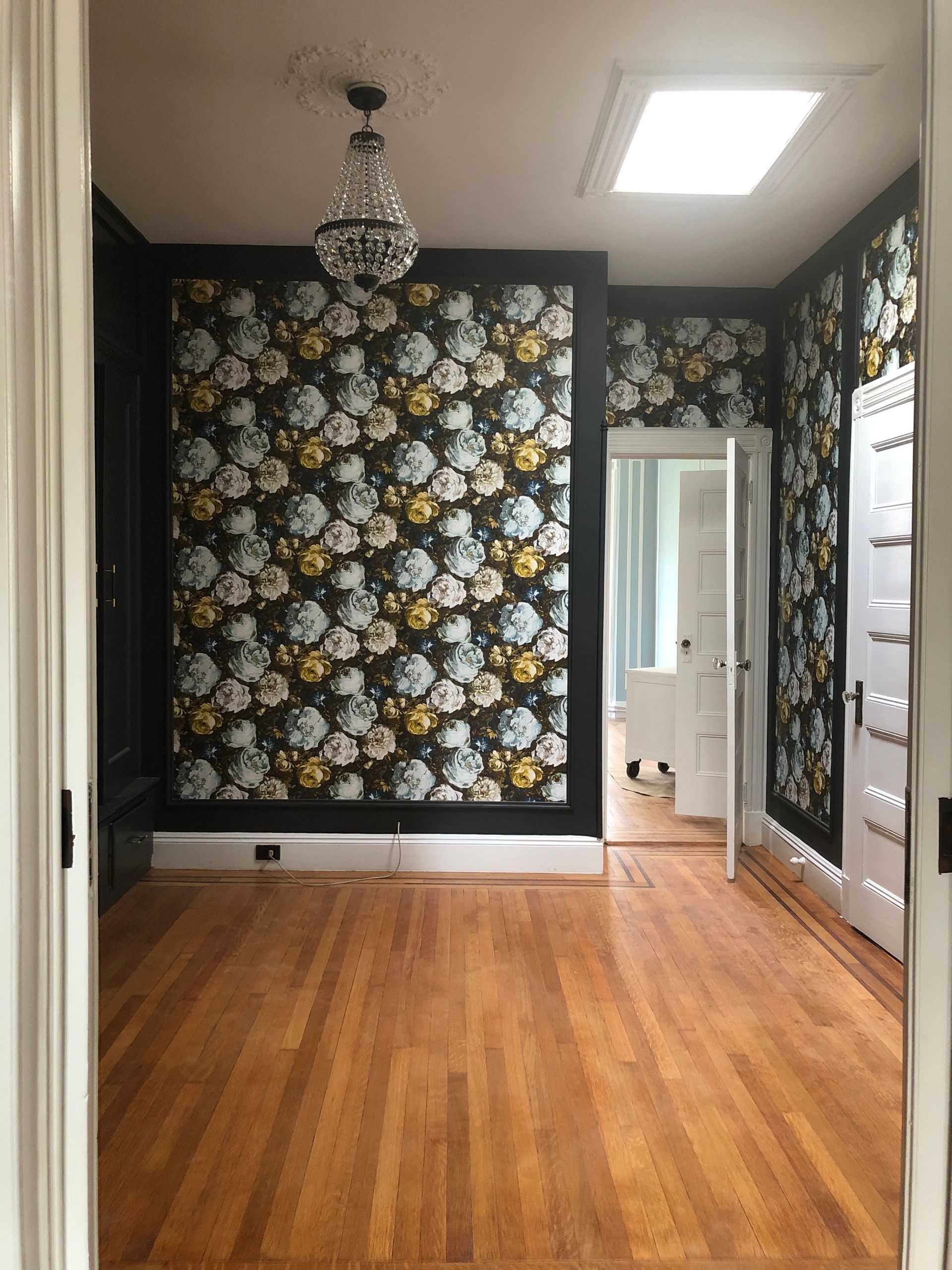 Empty room with wood floor, floral wallpaper, chandelier, and doorway to a bright adjoining space